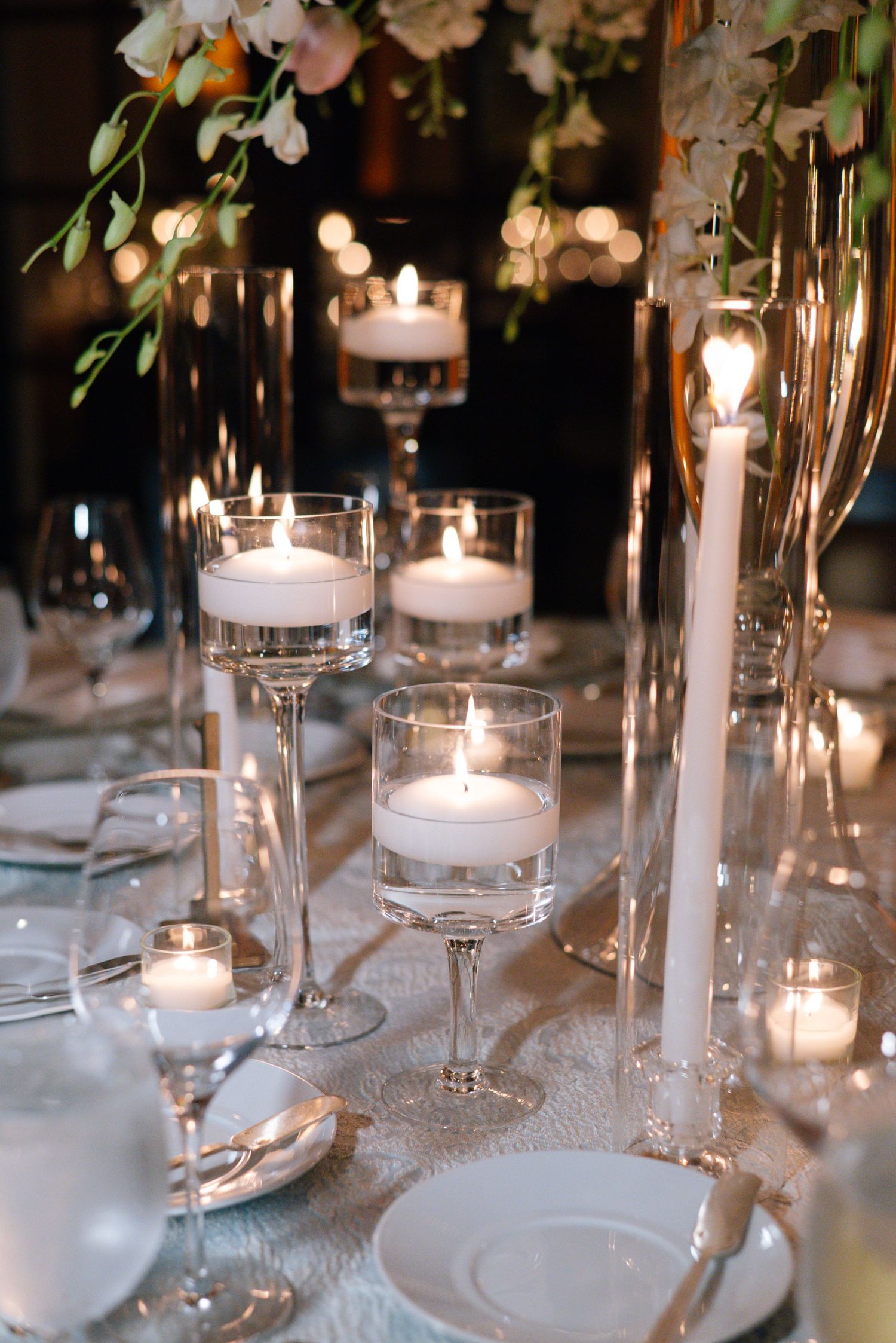 Elegant wedding table setting with floating candles in stemmed glass holders and soft ambient lighting.