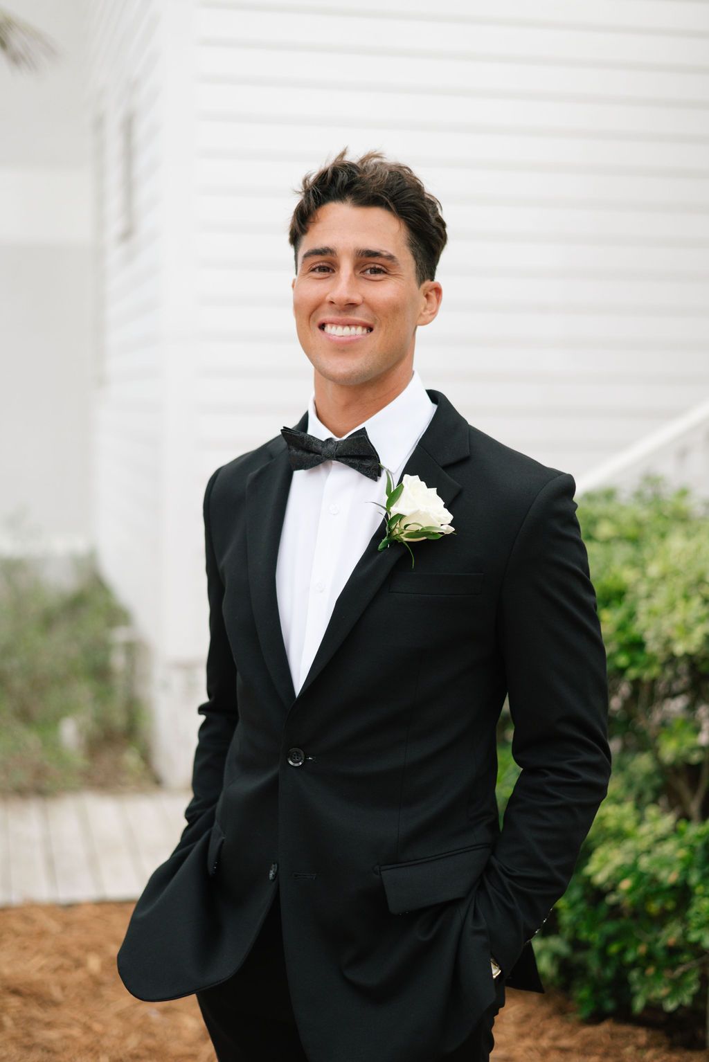 Groom in classic black tuxedo and bowtie poses against white building.