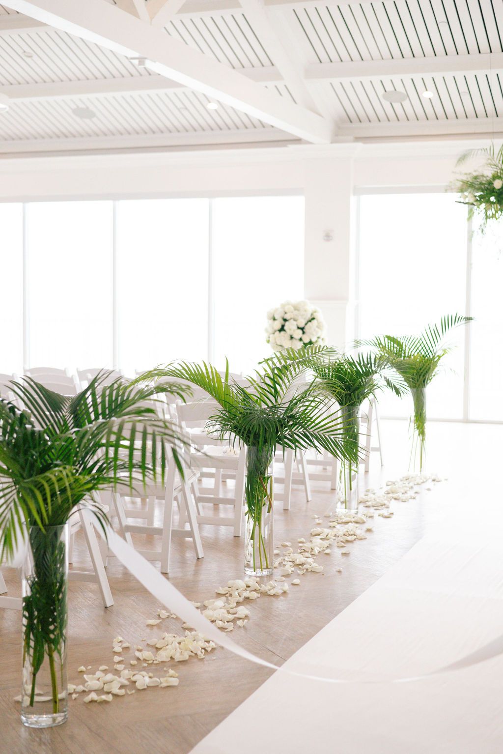 Light-filled wedding ceremony space with white decor, tropical plants, and floor-to-ceiling windows.