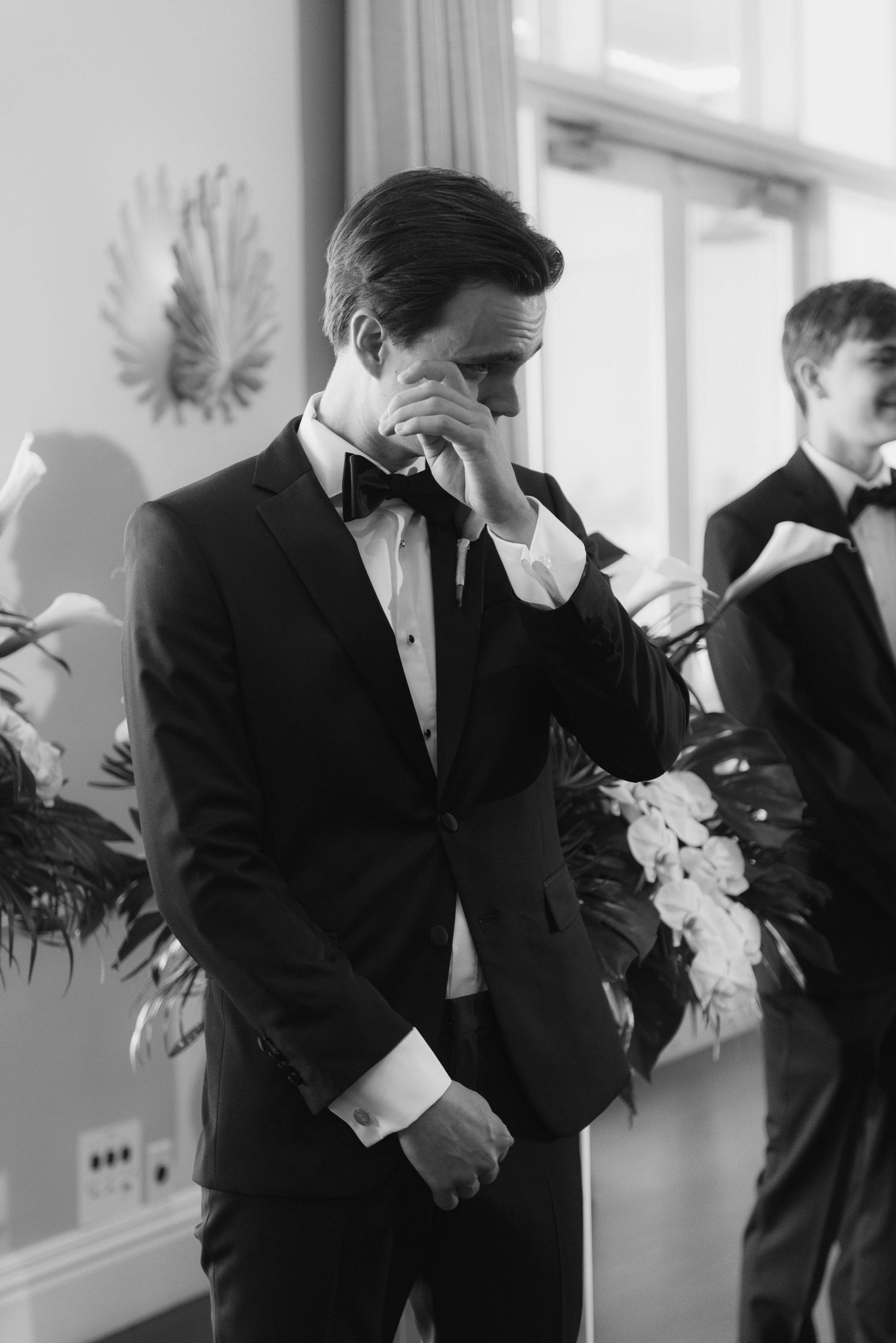 Wedding party members in black tuxedos prepare for ceremony indoors.