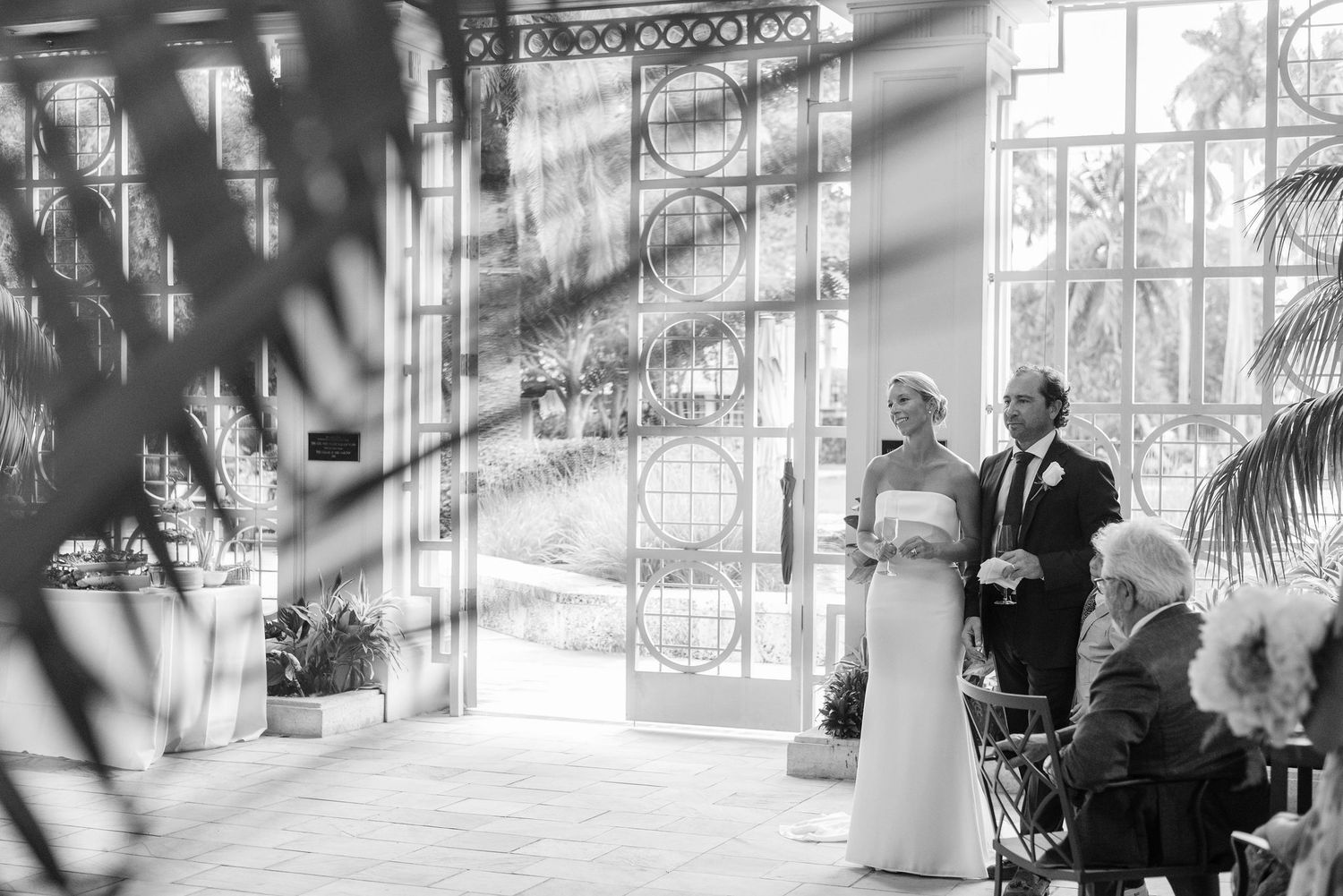 Black and white wedding ceremony photo in an elegant conservatory with palm trees and ornate windows.
