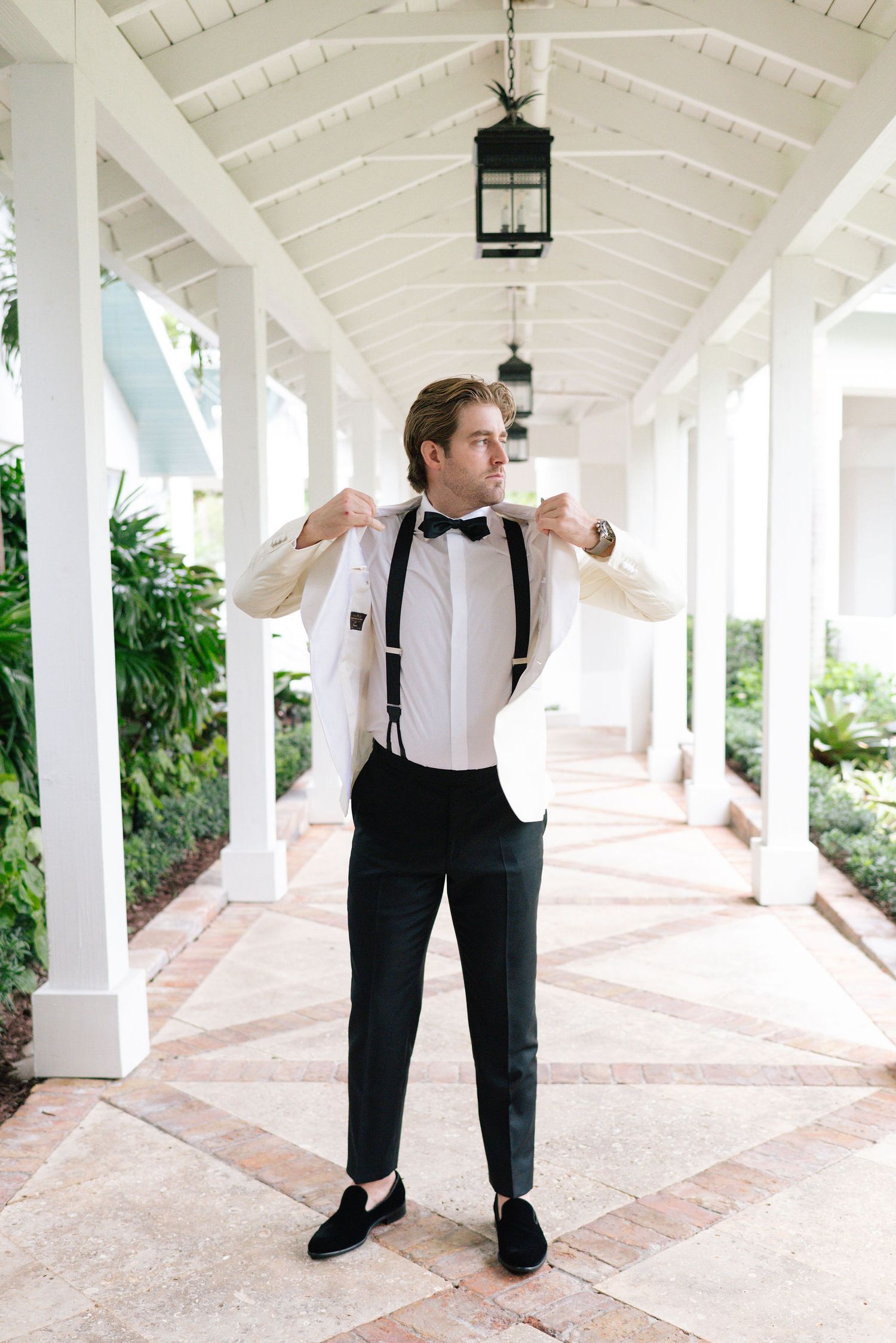Groom adjusts bowtie while standing in white columned corridor wearing classic tuxedo with suspenders.