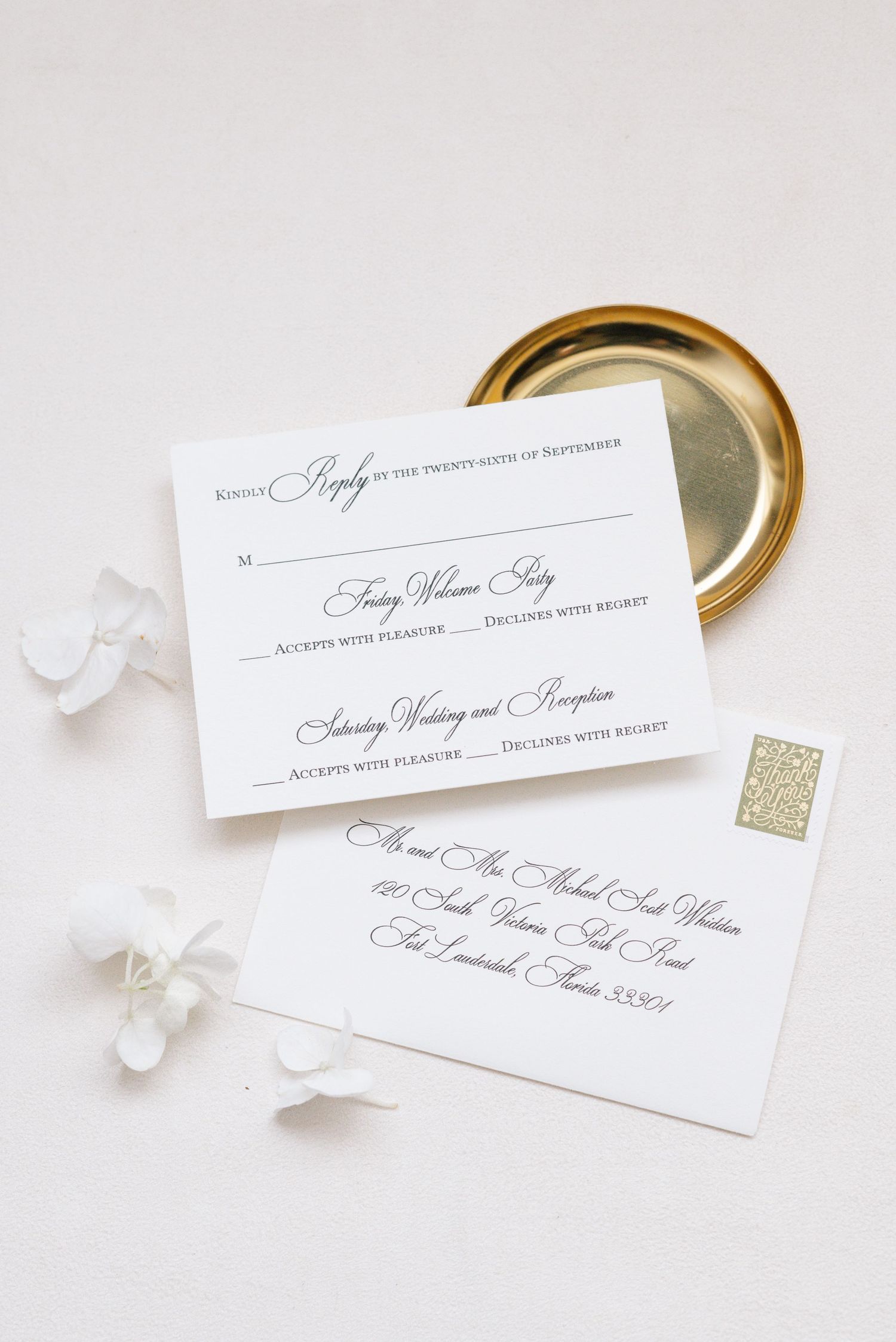 Classic white wedding RSVP card with cursive script beside gold dish and white flowers.
