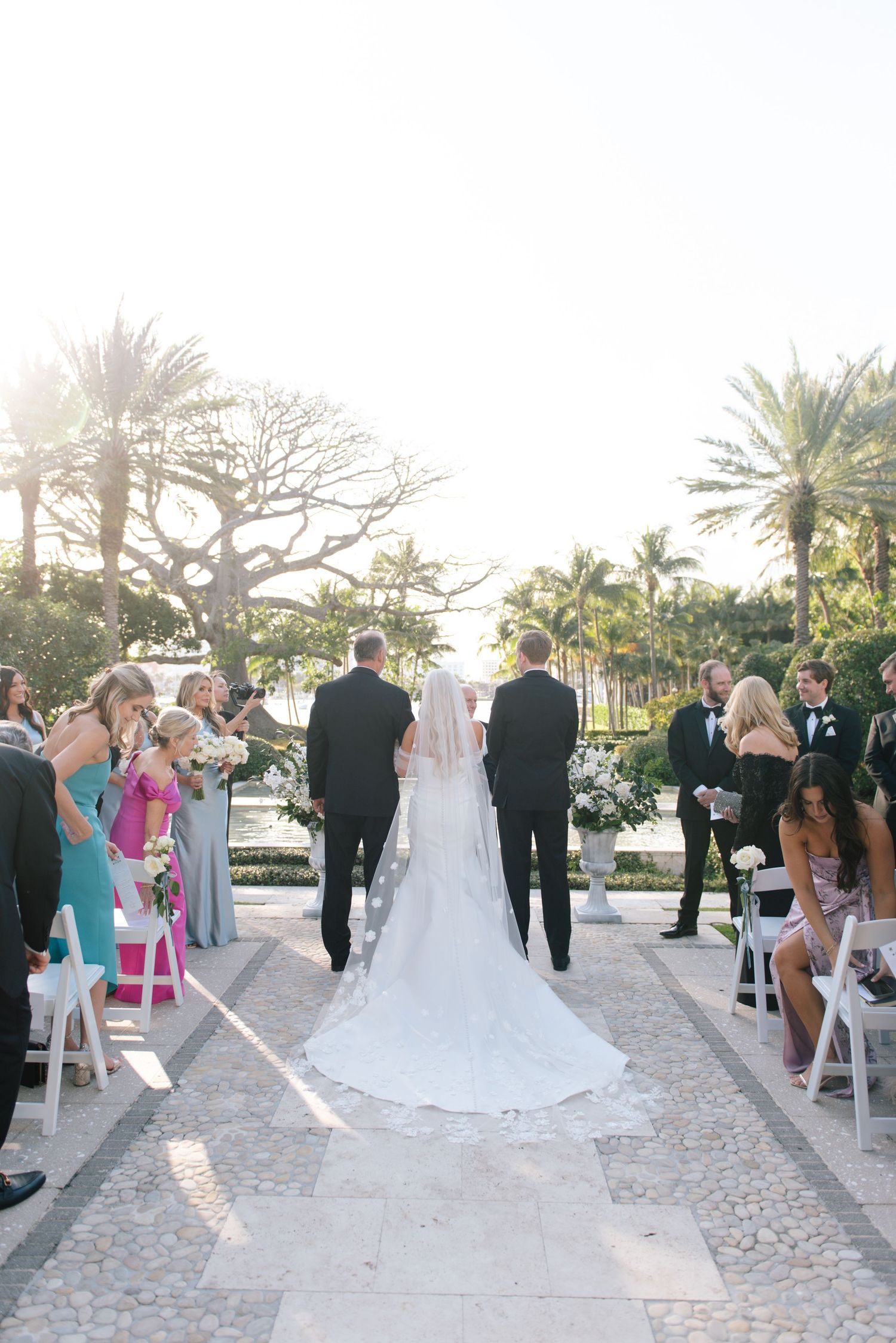 A bride walks down an outdoor aisle with her long white veil trailing behind at a tropical palm-lined venue.