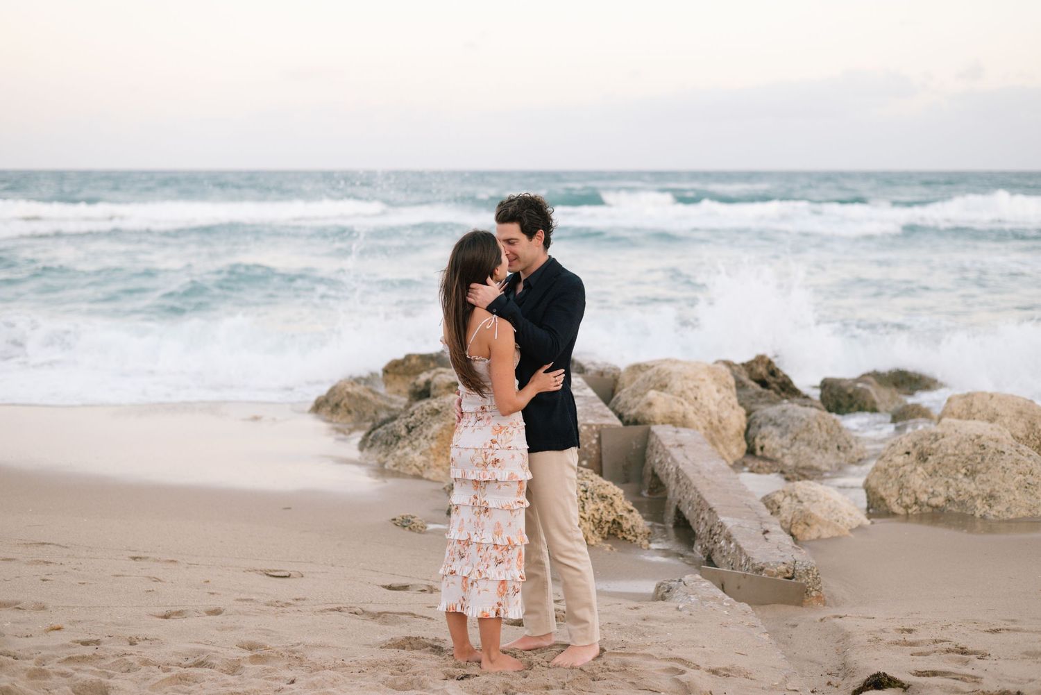 A couple embraces on a sandy beach with waves and rocky shoreline during golden hour.
