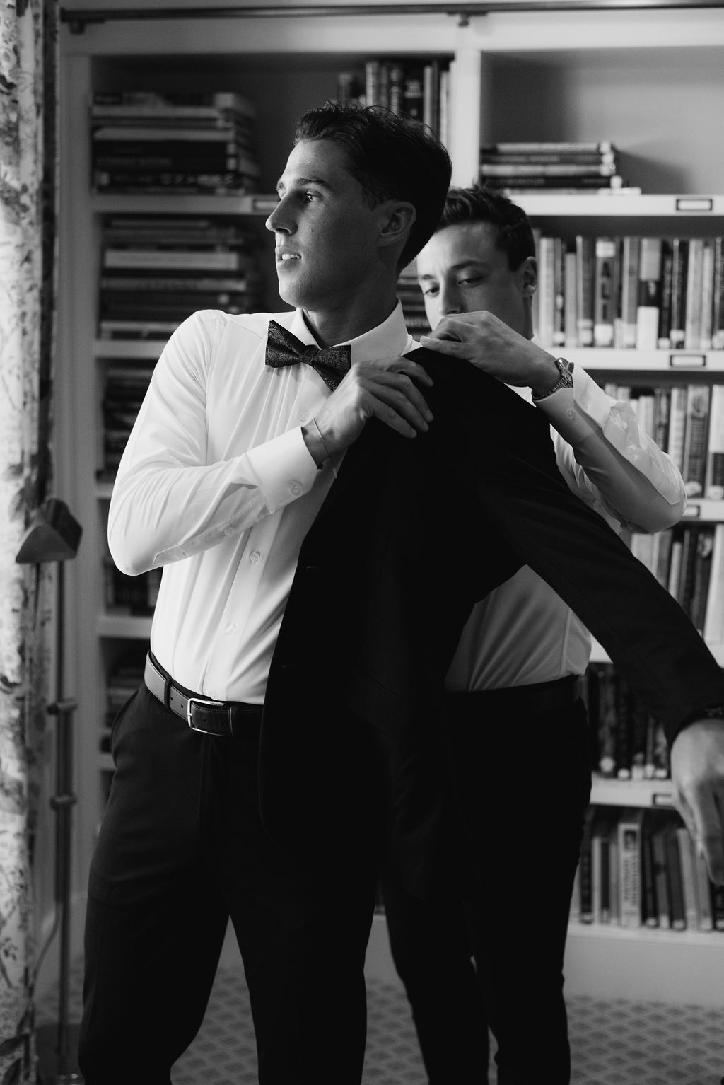 Two people in formal attire helping each other get dressed in front of a bookshelf.