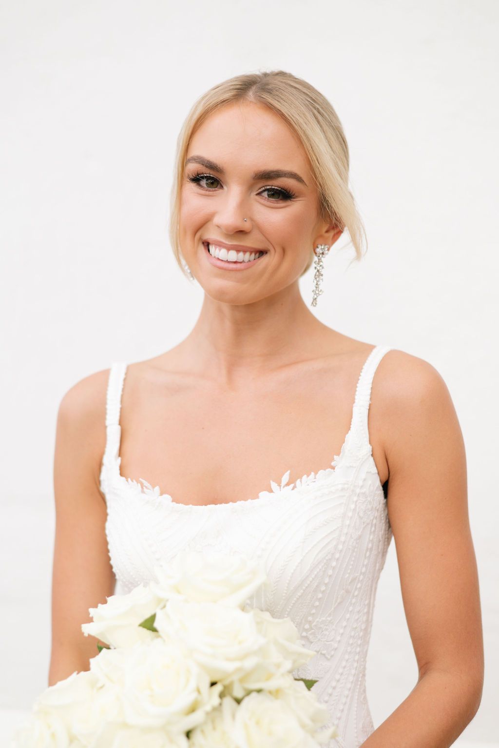 Bride in white sequined dress holding elegant white bouquet against light background.