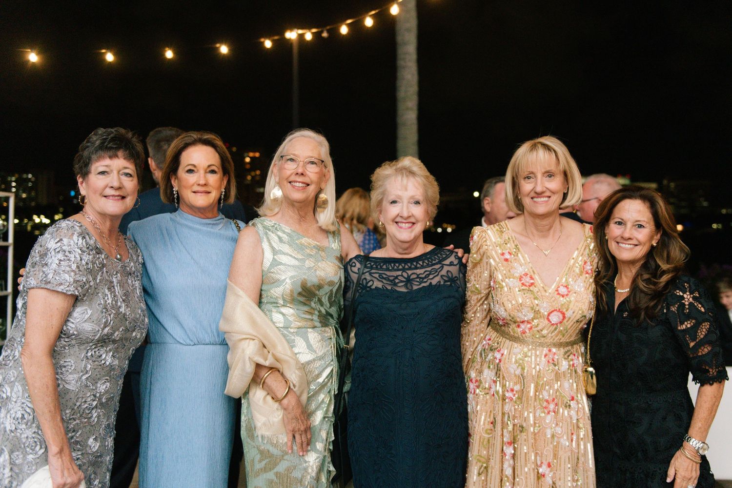 Six elegantly dressed women smile together at outdoor evening wedding reception.