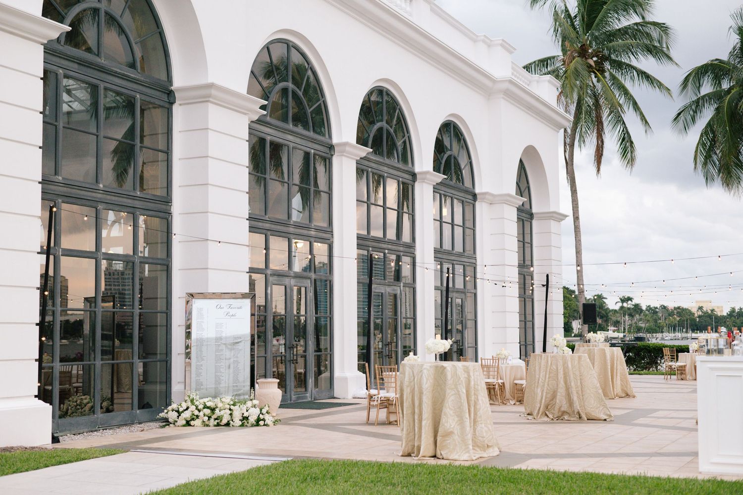 Elegant outdoor wedding reception venue with white arched windows, palm trees, and draped cocktail tables.