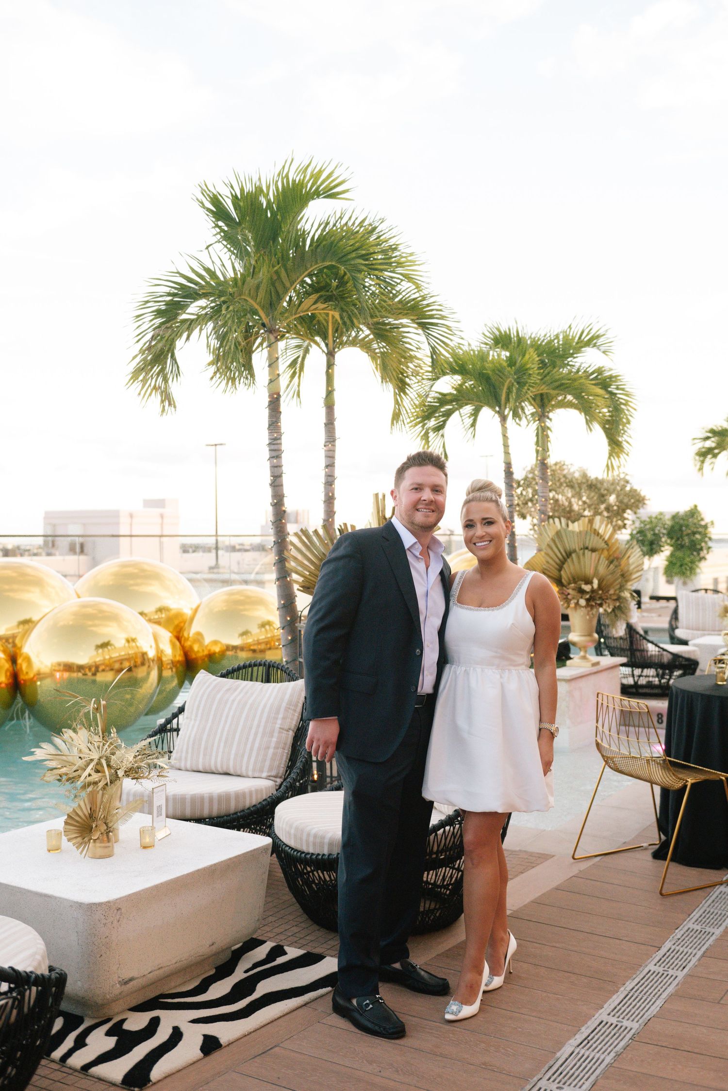 Elegant outdoor event setup with white furniture and palm trees against a sunset backdrop.
