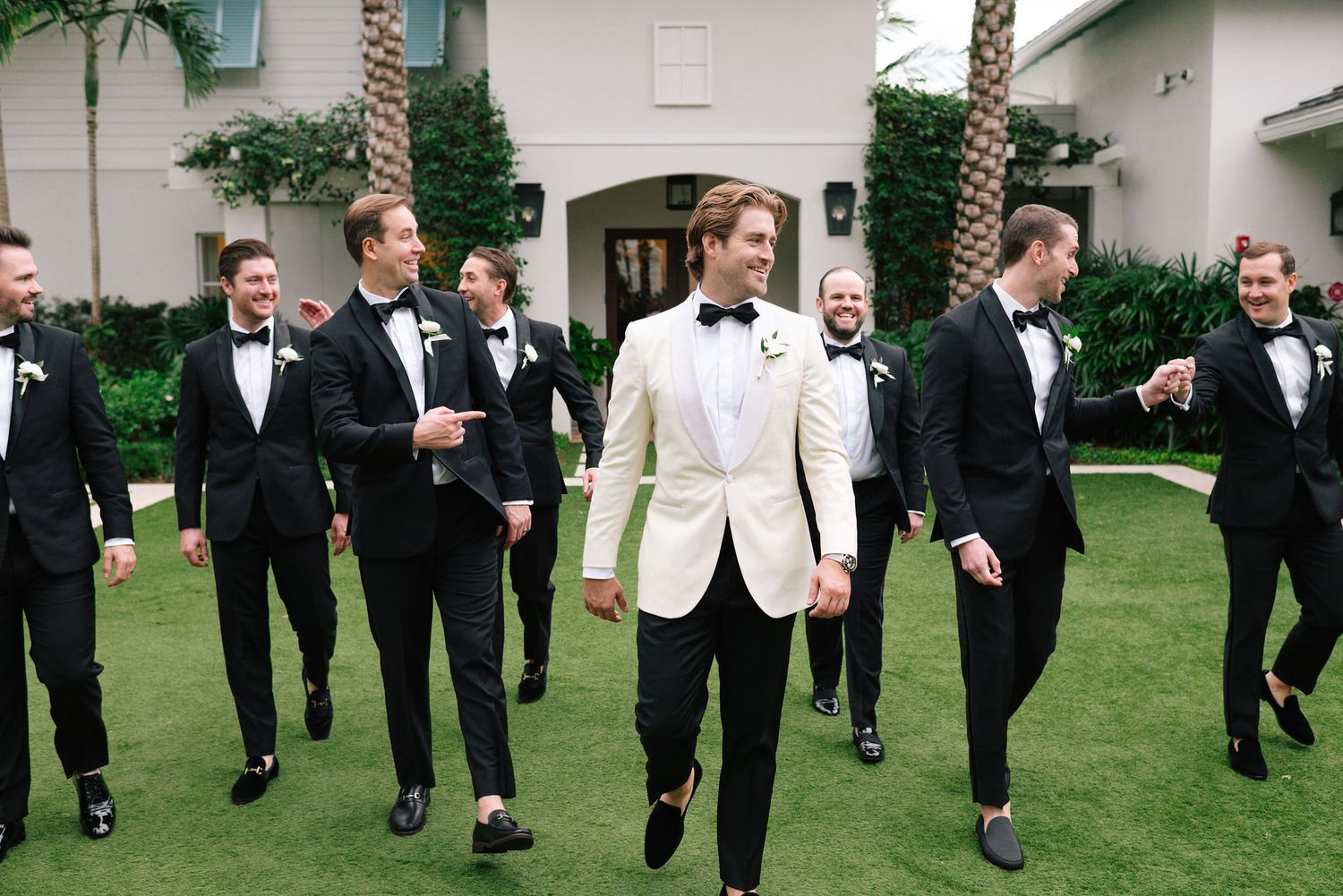 Groomsmen in black suits walking and laughing on green lawn.