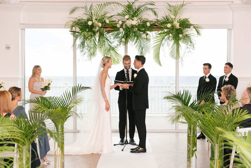 Intimate wedding ceremony in white coastal venue with tropical greenery decorations.