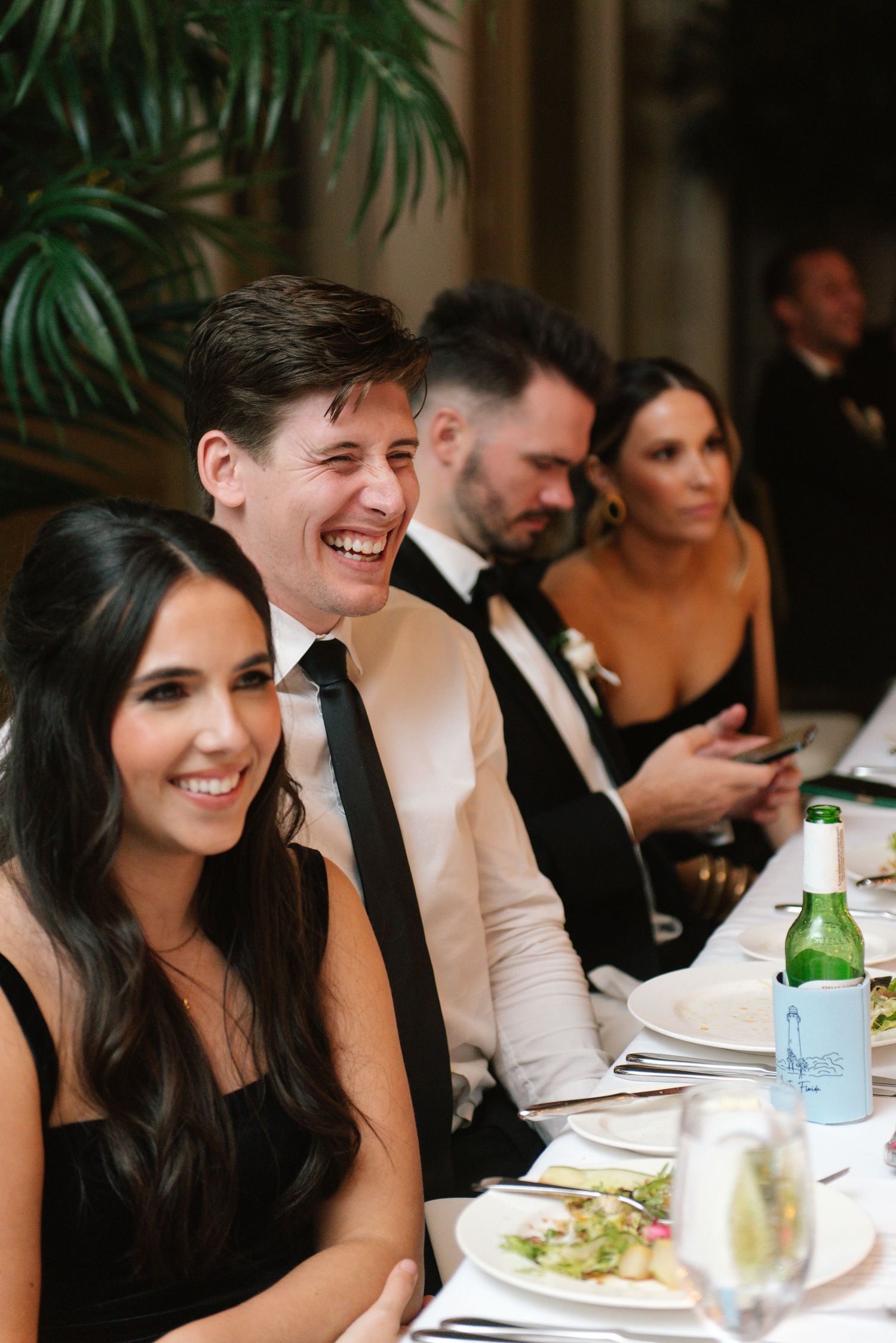 Wedding party members smile and laugh while seated at an elegant reception dinner table.