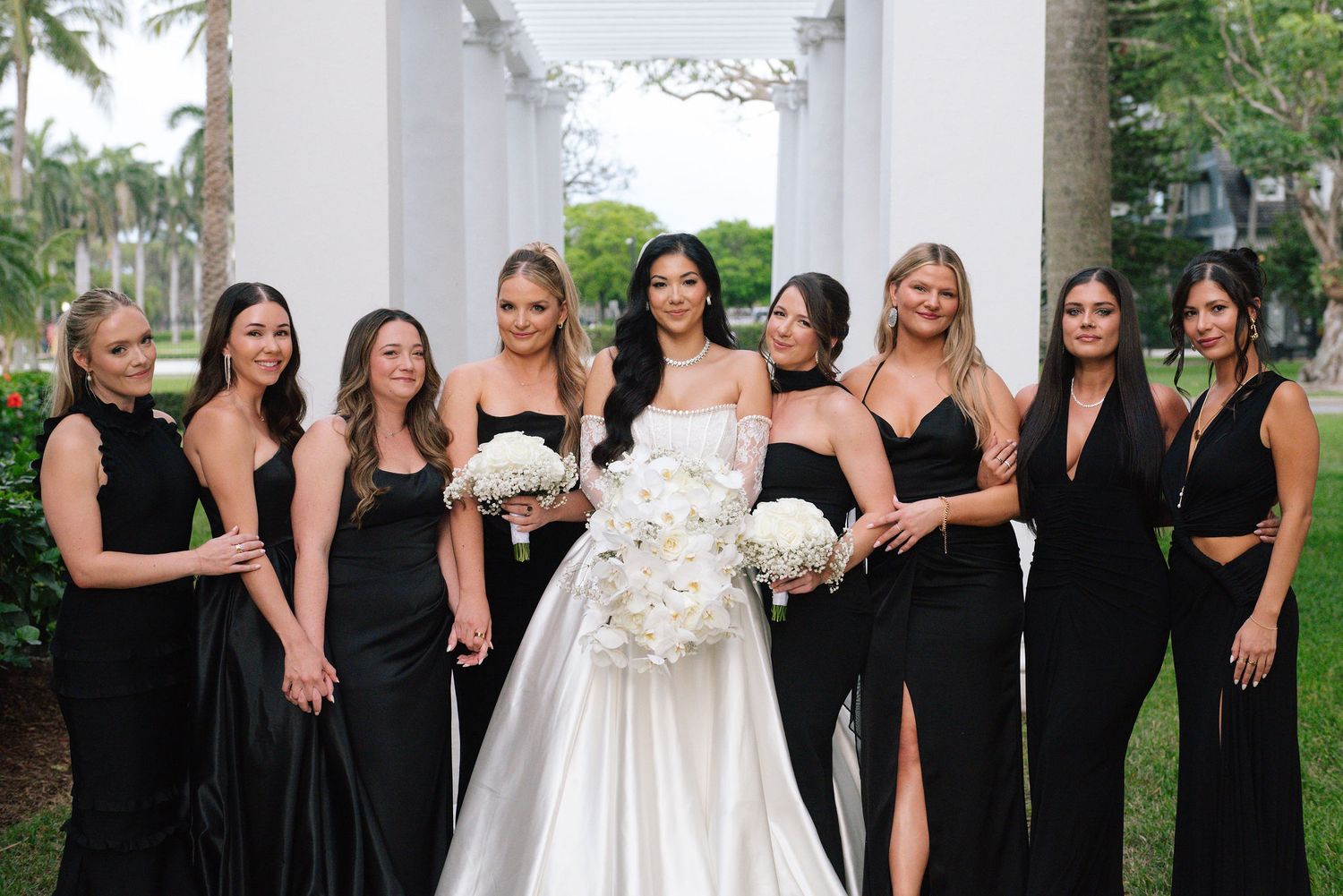 Bridesmaids in black dresses pose together outdoors near white columns.