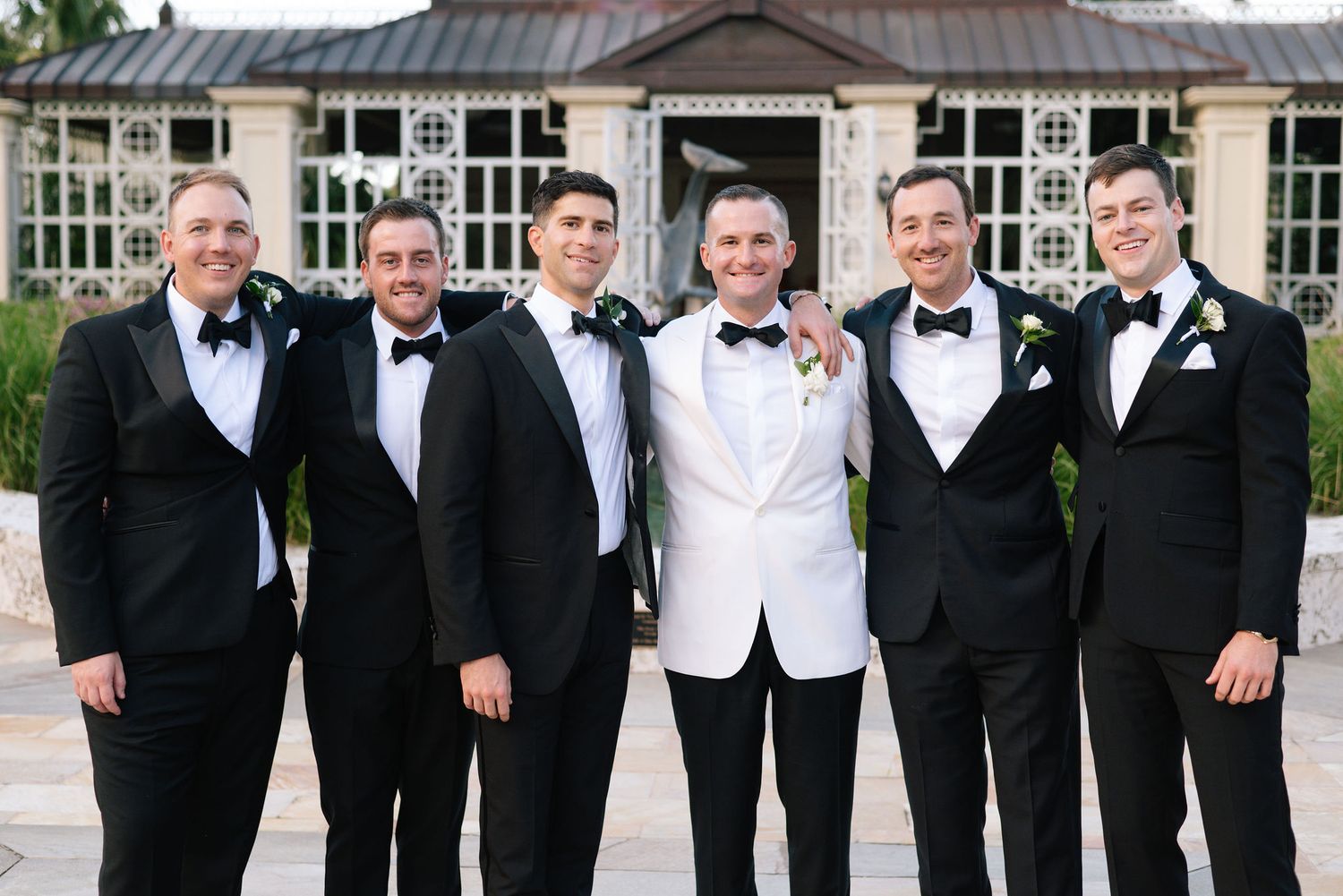 A wedding party wearing black tuxedos and bow ties pose together in formal attire.