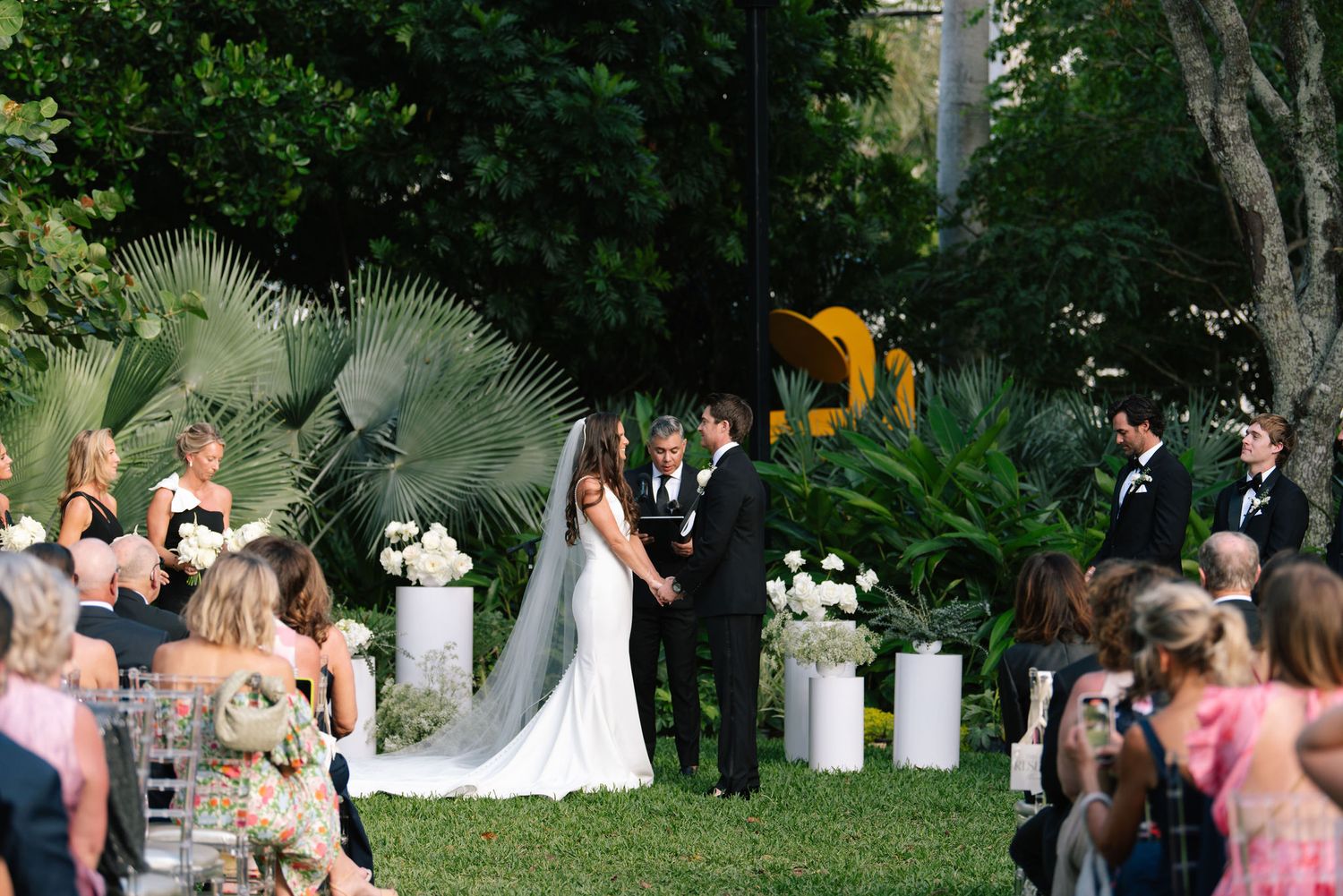Outdoor wedding ceremony in a lush garden setting with palm trees and white floral decorations.