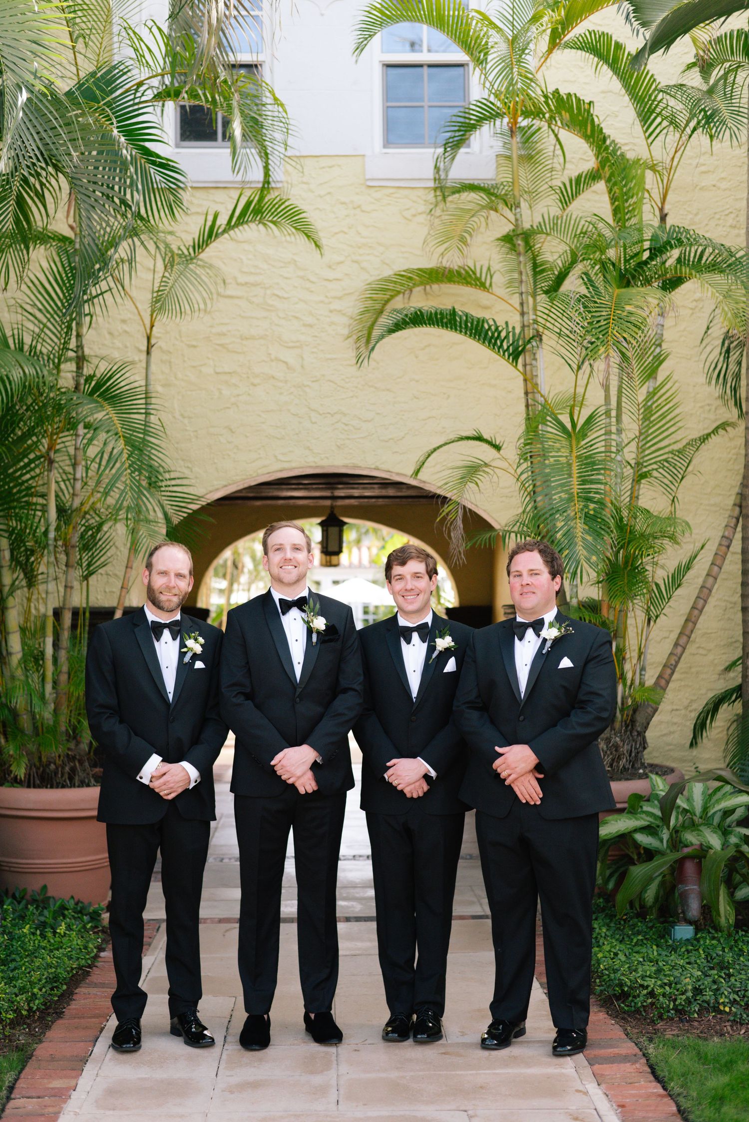 Four groomsmen in black tuxedos stand under an archway surrounded by tropical palm trees.