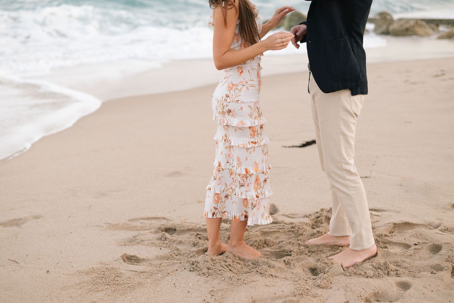 Two pairs of feet stand close together on sandy beach with waves in background and floral dress visible.