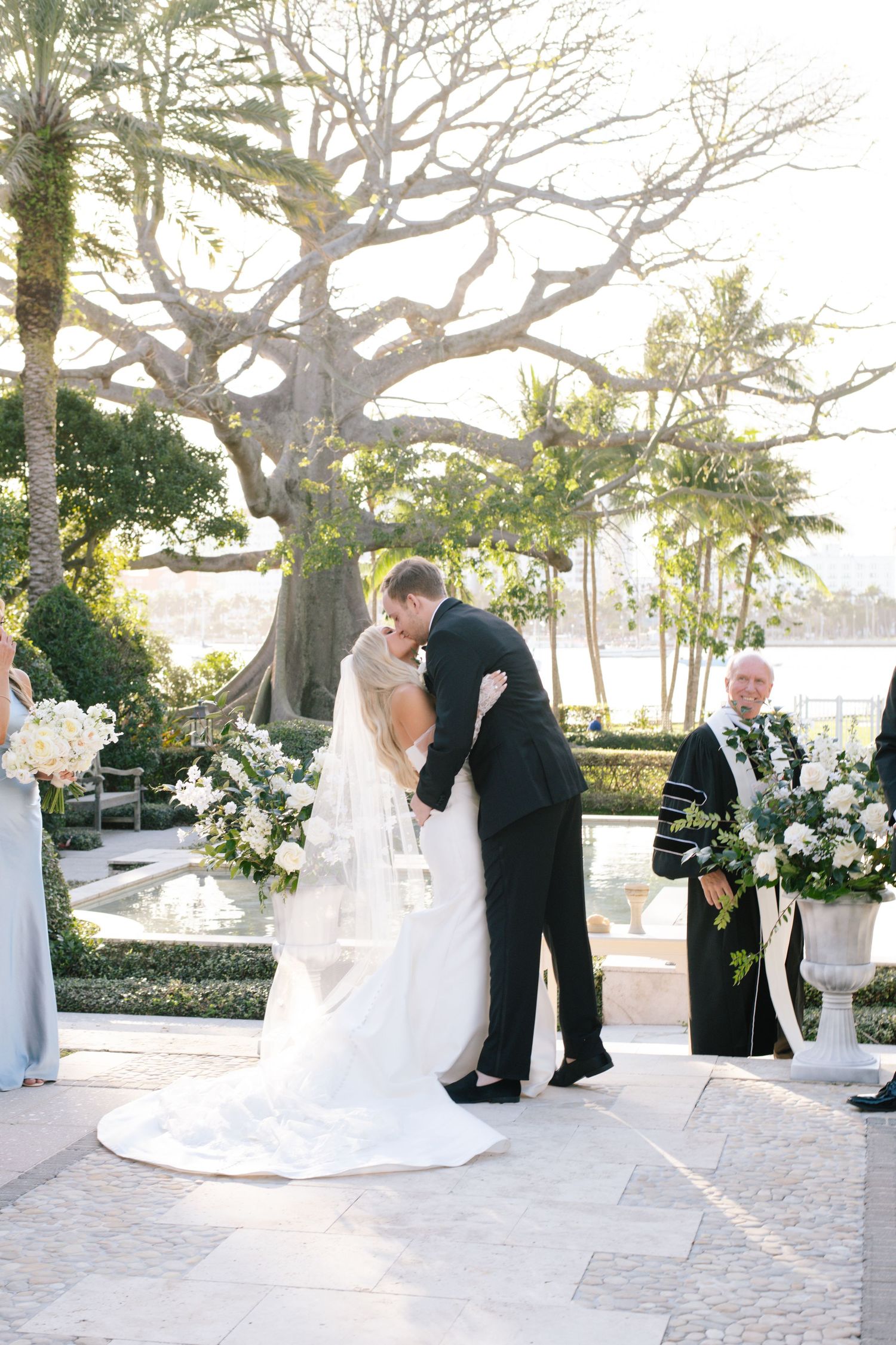 A wedding ceremony takes place under a large tree with white floral arrangements and waterfront views.