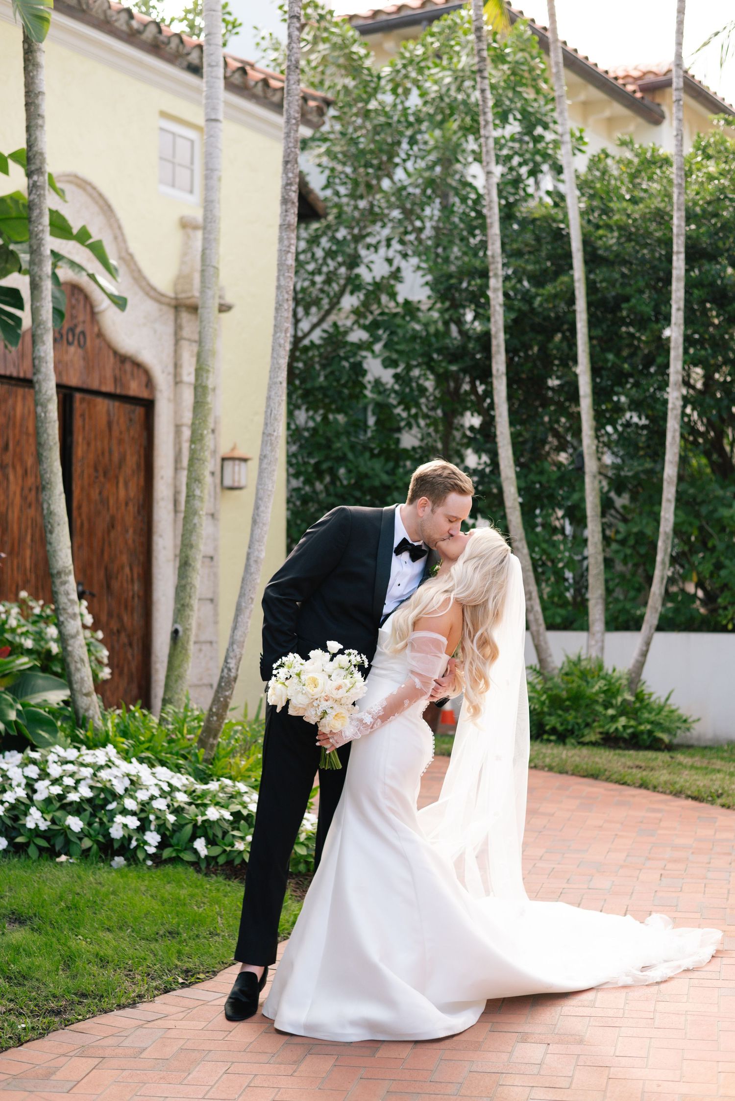 Wedding couple shares romantic moments on a brick patio surrounded by white flowers.