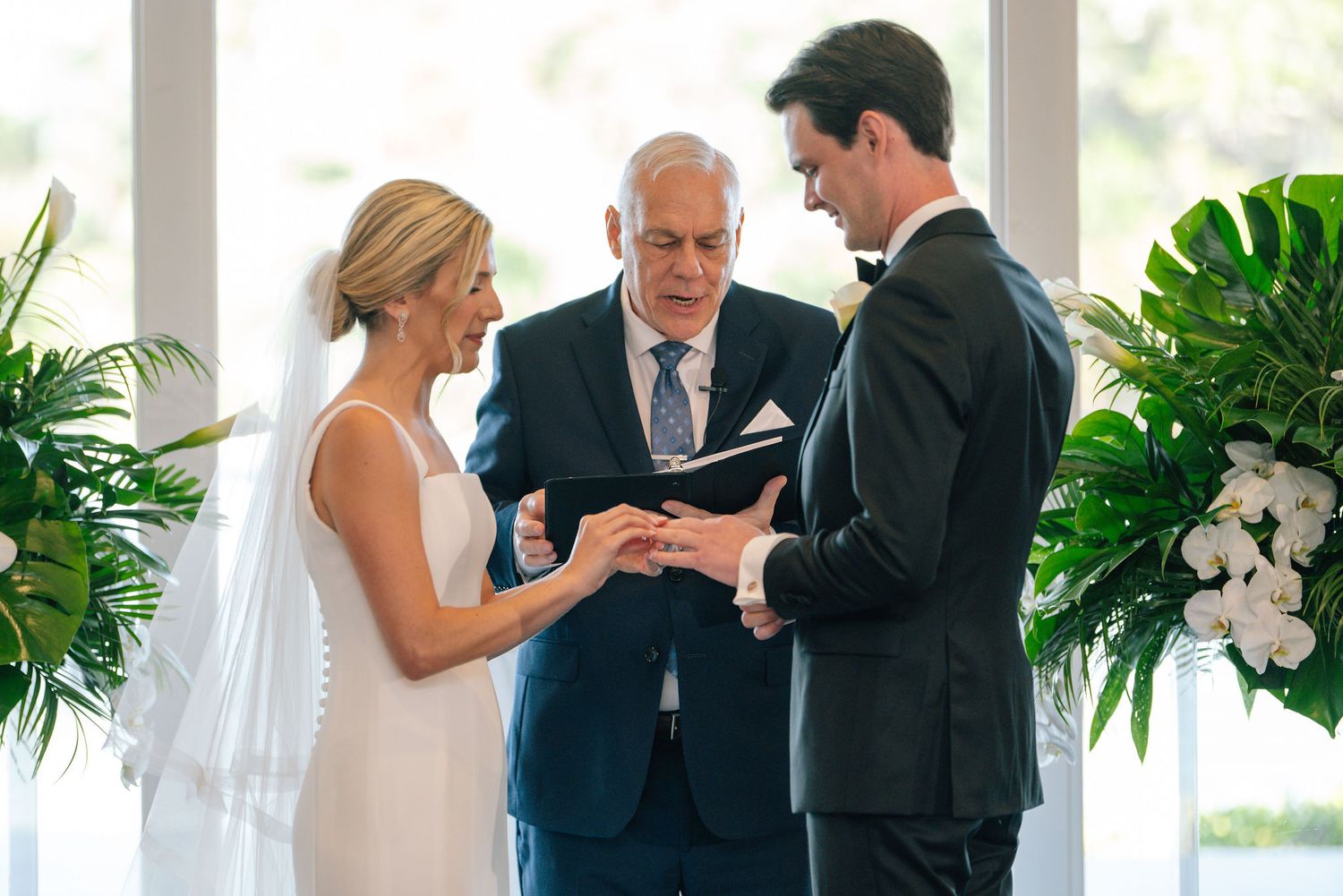 Wedding ceremony exchange of rings with officiant in bright sunlit venue.