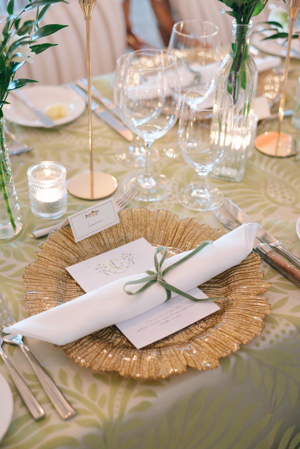 Place setting featuring gold charger plates, white napkins, and green botanical accents.