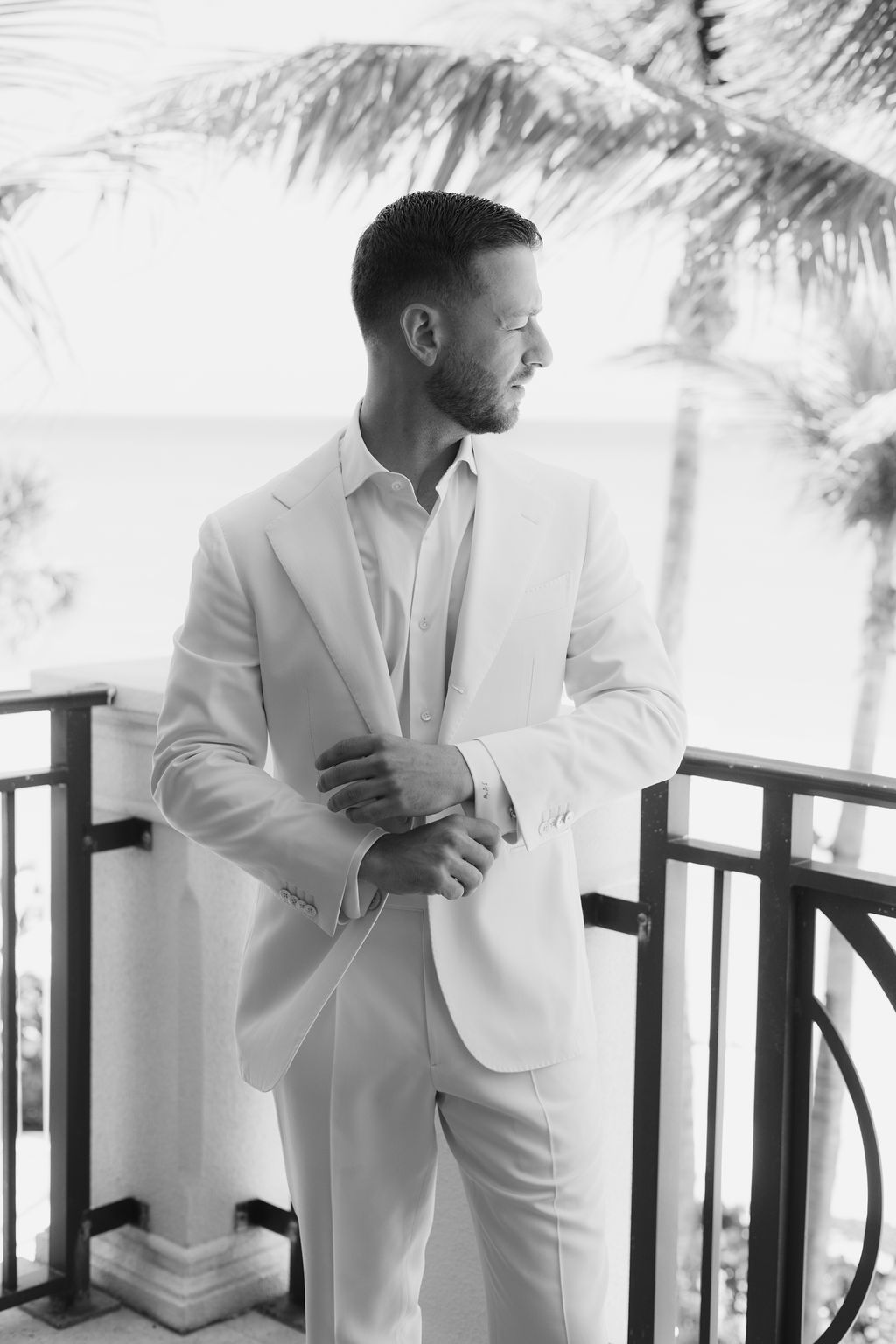 Groom in white suit adjusting cuffs near tropical palm trees.