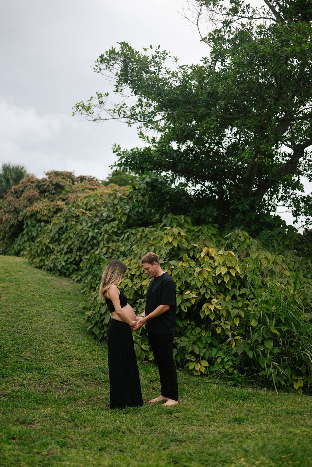 Couple shares tender moments while standing on a grassy hill with trees in the background.