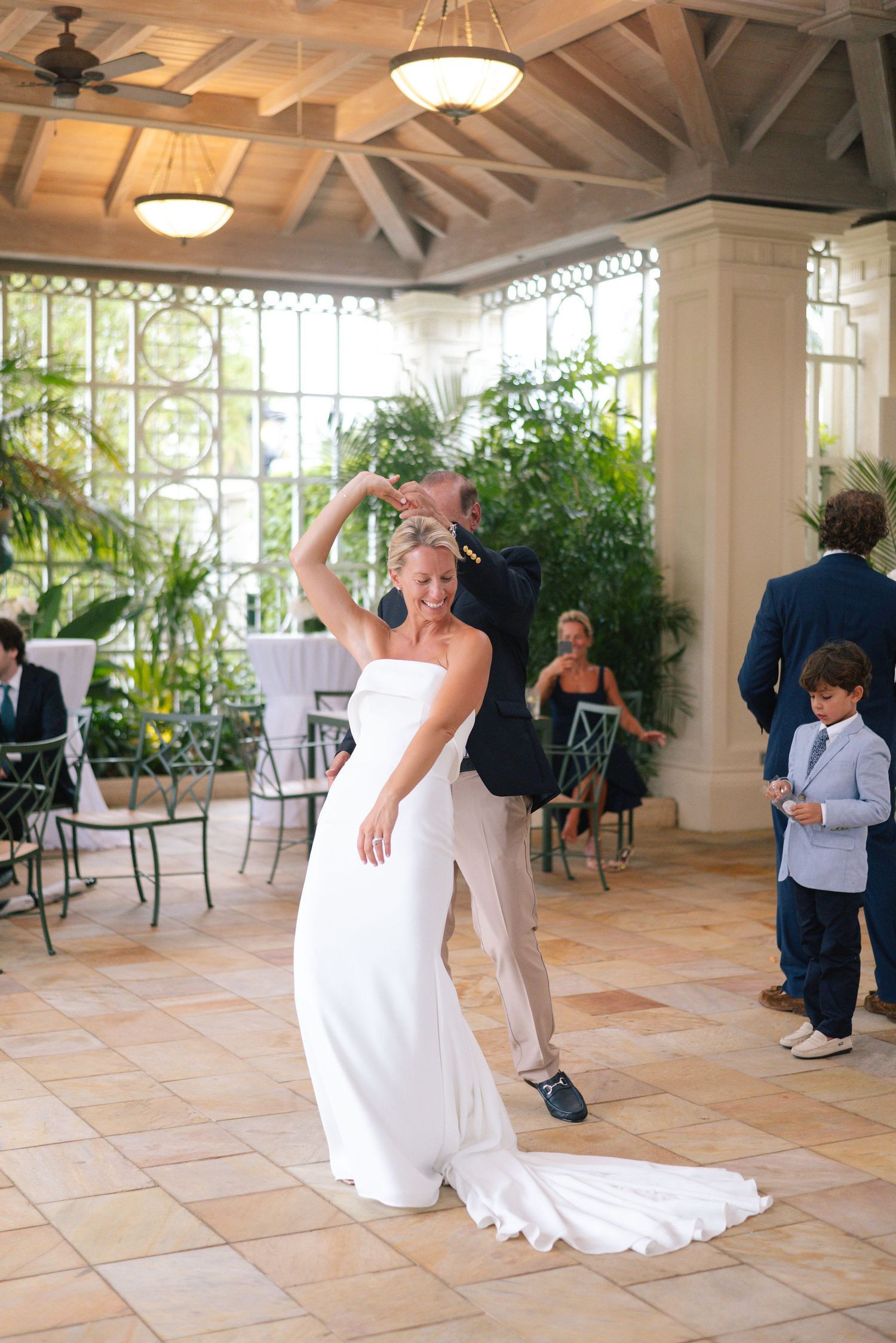 Bride in flowing white gown dancing solo in a sunlit conservatory space.