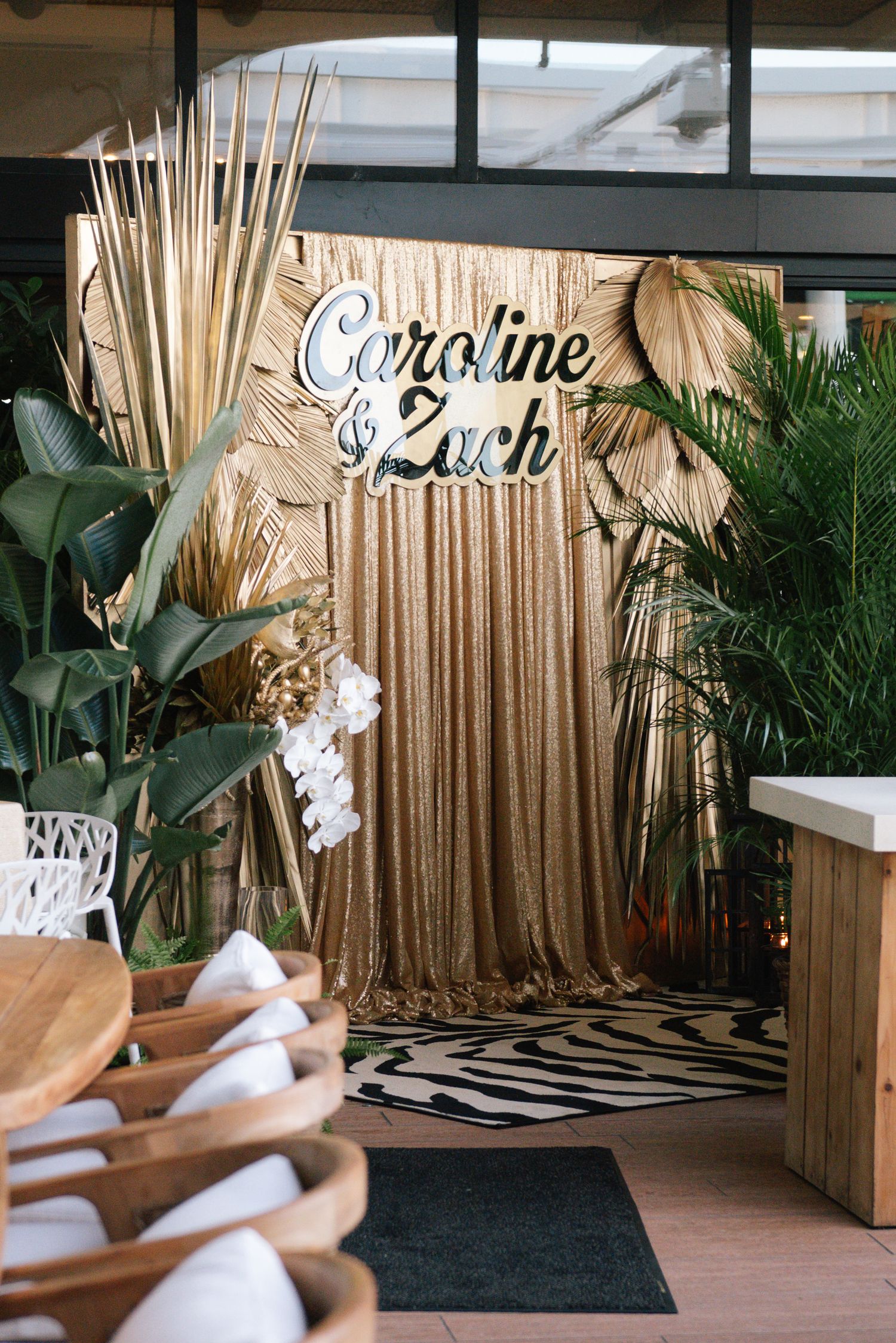 Elegant wedding backdrop with bamboo decor, tropical plants, and custom name signage against a gold curtain.