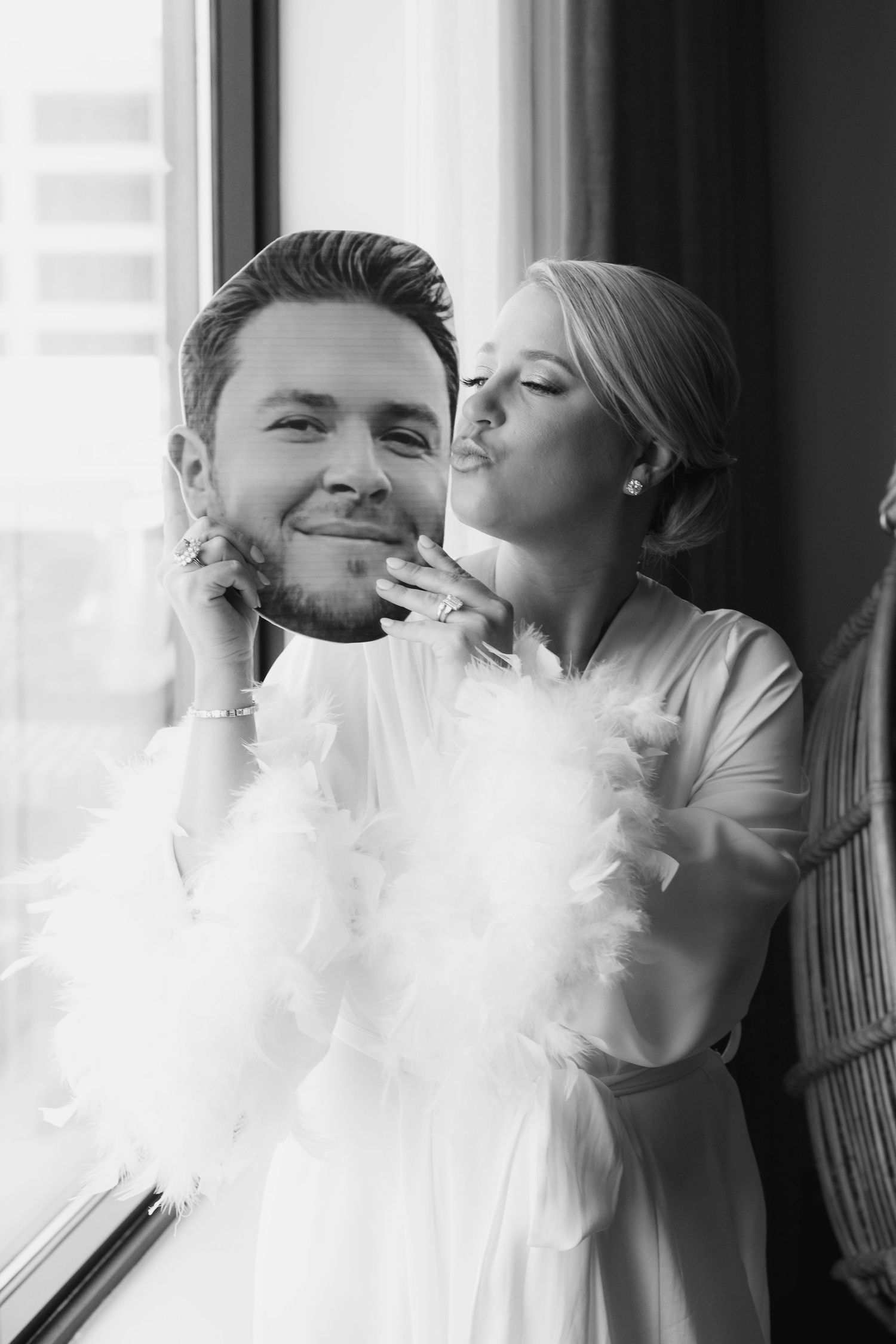 Bride kisses a cutout photo of groom while wearing a white feather boa in a black and white wedding photo.