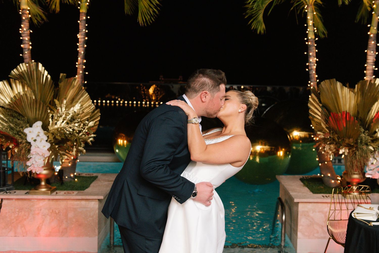 Romantic nighttime kiss between newlyweds with palm trees and string lights in background.