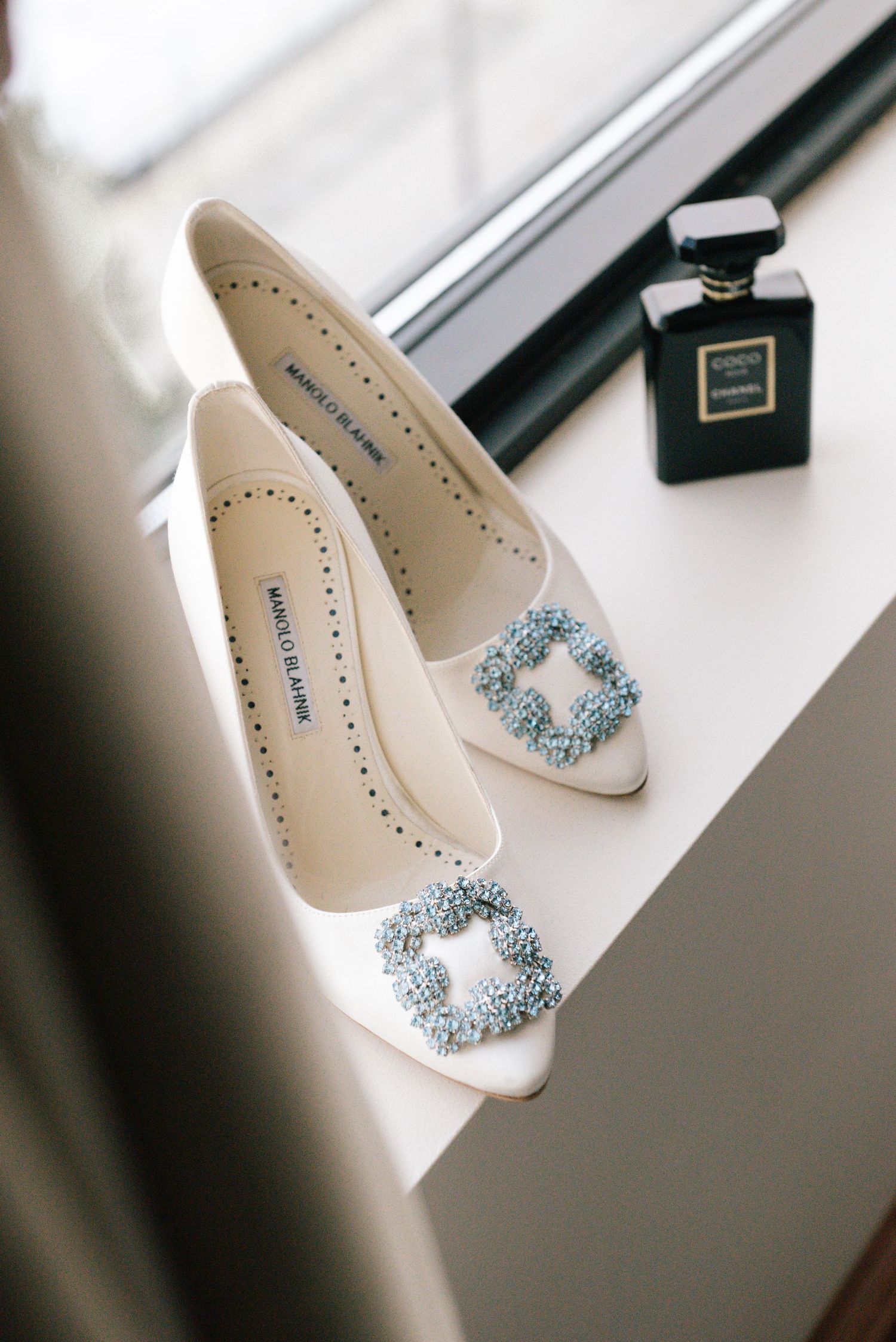 Manolo Blahnik white satin heels with blue crystal buckles beside a Chanel perfume bottle on a white windowsill.