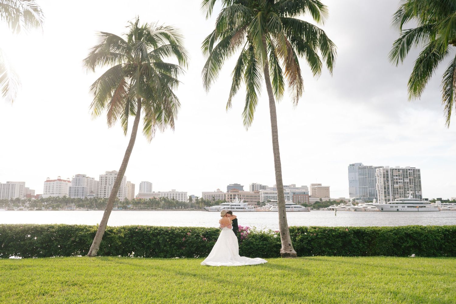 Bride and groom embracing between palm trees with waterfront city skyline backdrop.