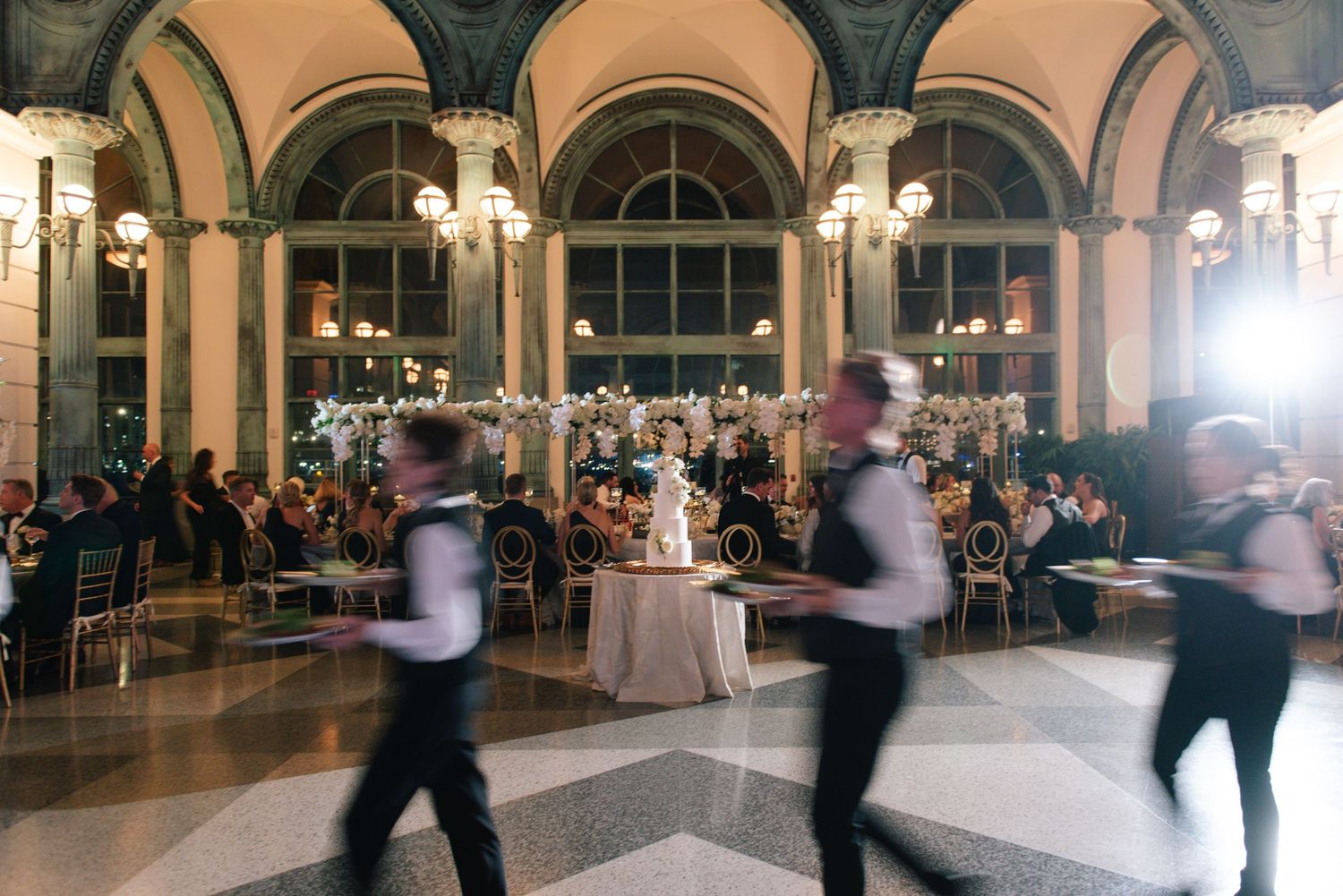 Waiters moving through elegant ballroom wedding reception with tall floral centerpieces.
