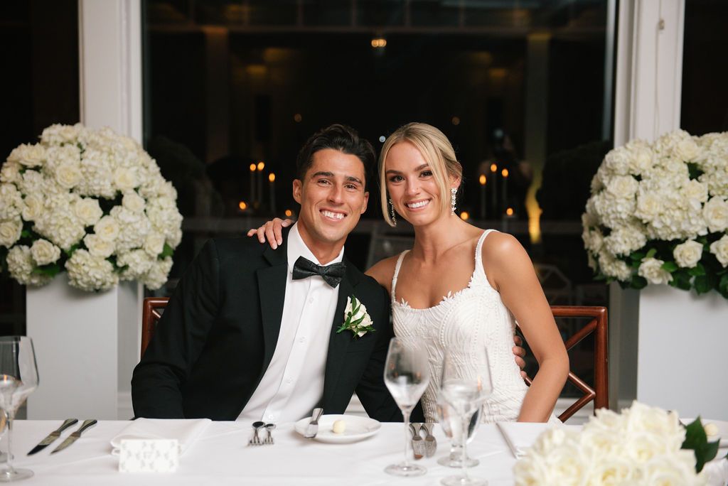Couple sits at elegant head table decorated with white floral arrangements.
