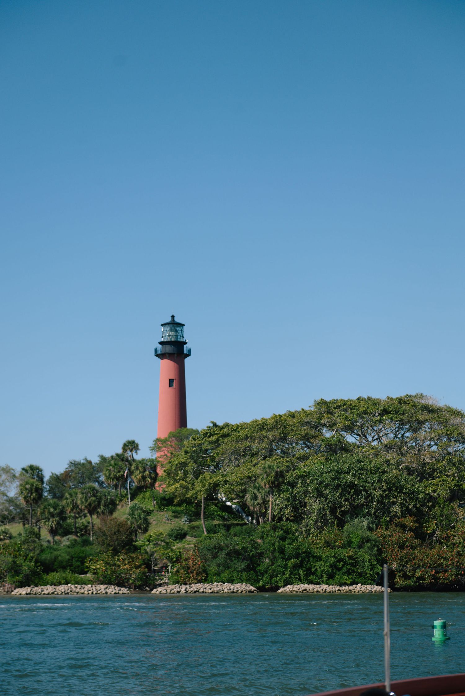 Red and white lighthouse stands tall against blue sky on tropical waterfront.