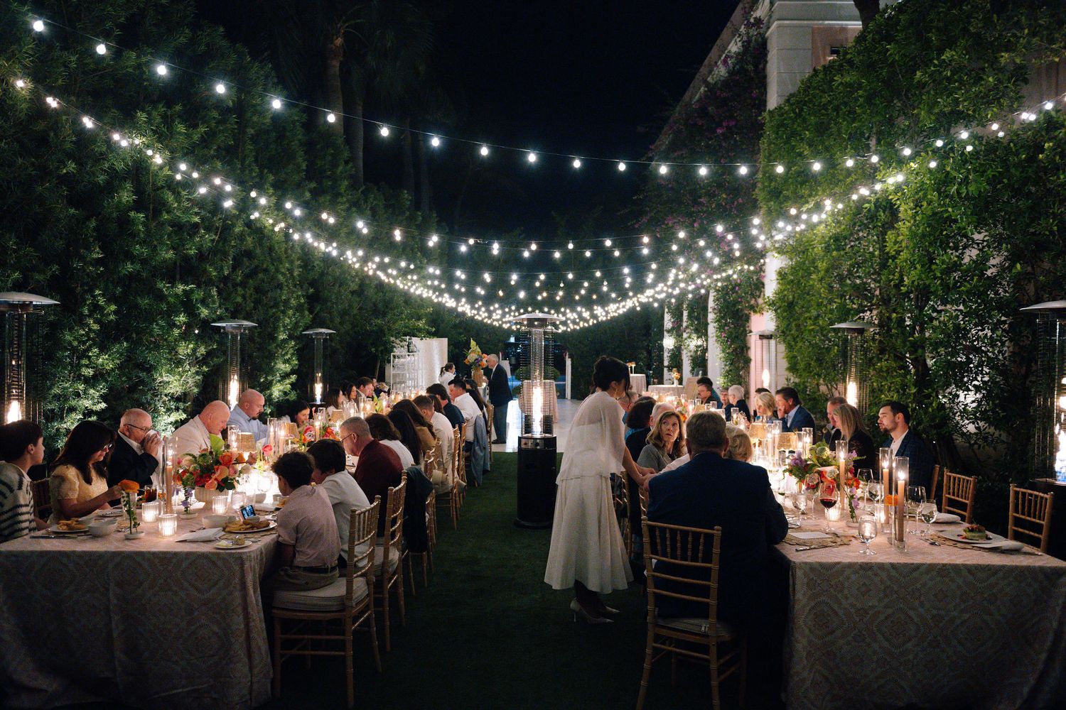 Outdoor wedding reception with string lights illuminating long tables under the night sky.
