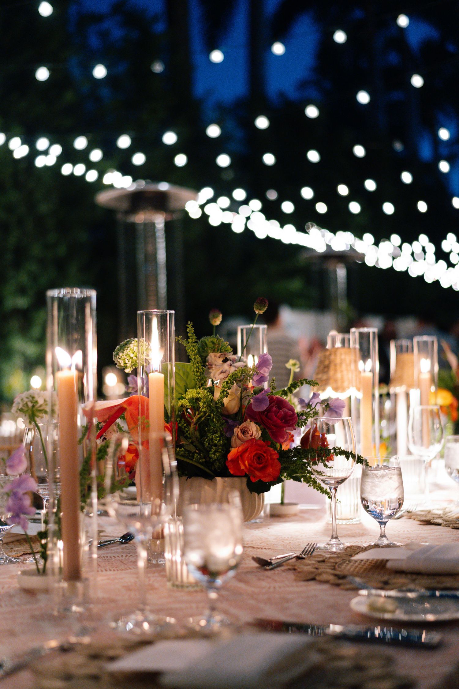 Candlelit outdoor dining table with glassware and floral centerpieces under string lights.