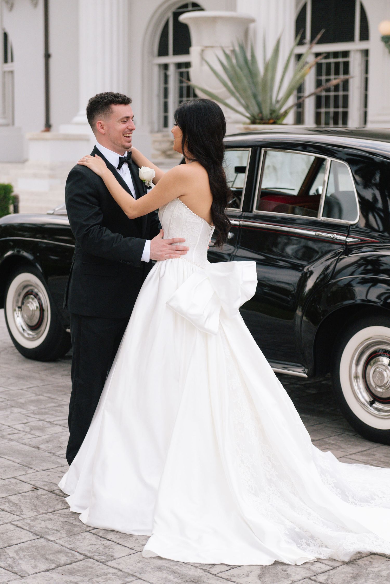 Wedding couple dances by classic black car outside elegant white building.