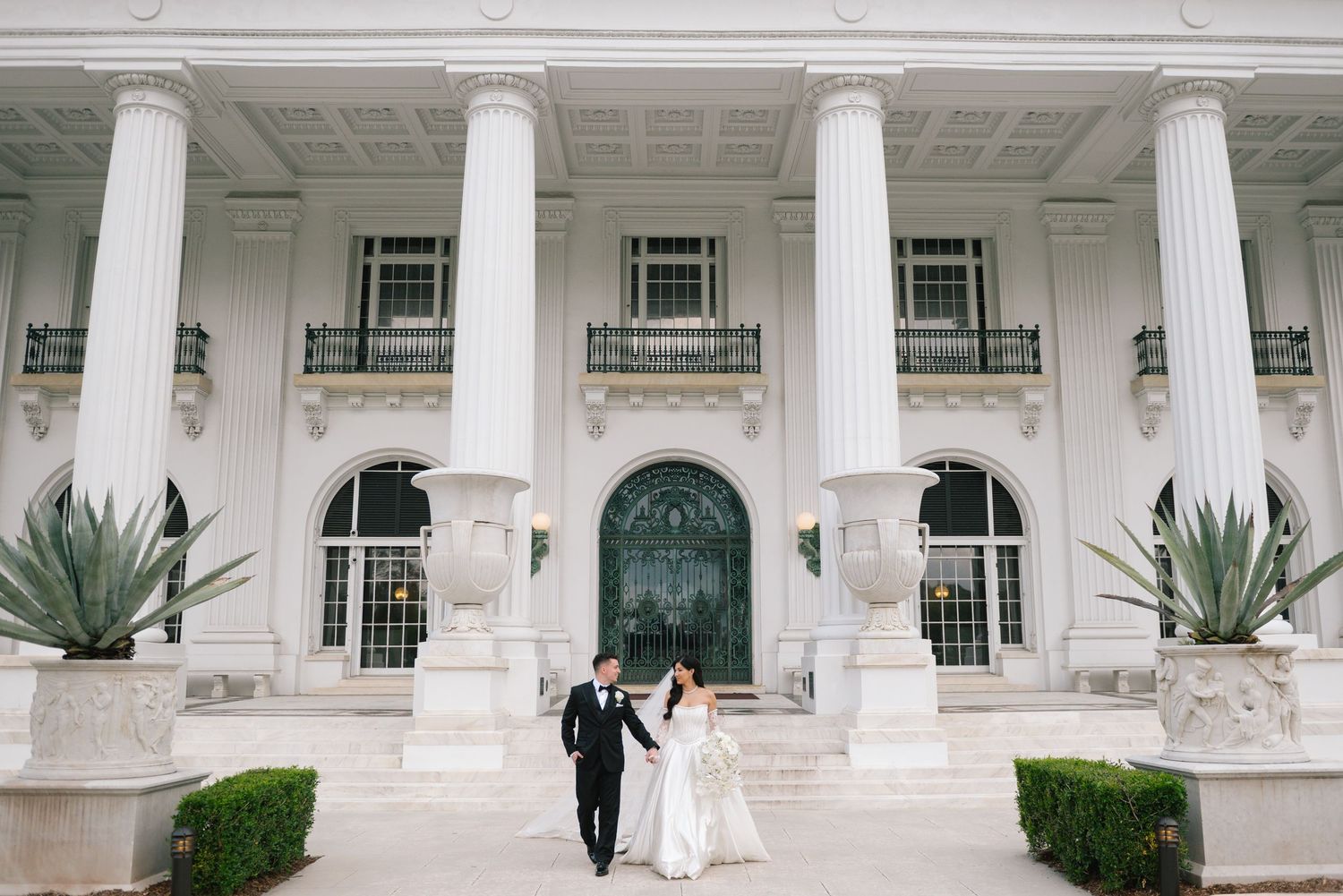 Elegant wedding couple stands before a grand white building with classical columns and ornate architecture.