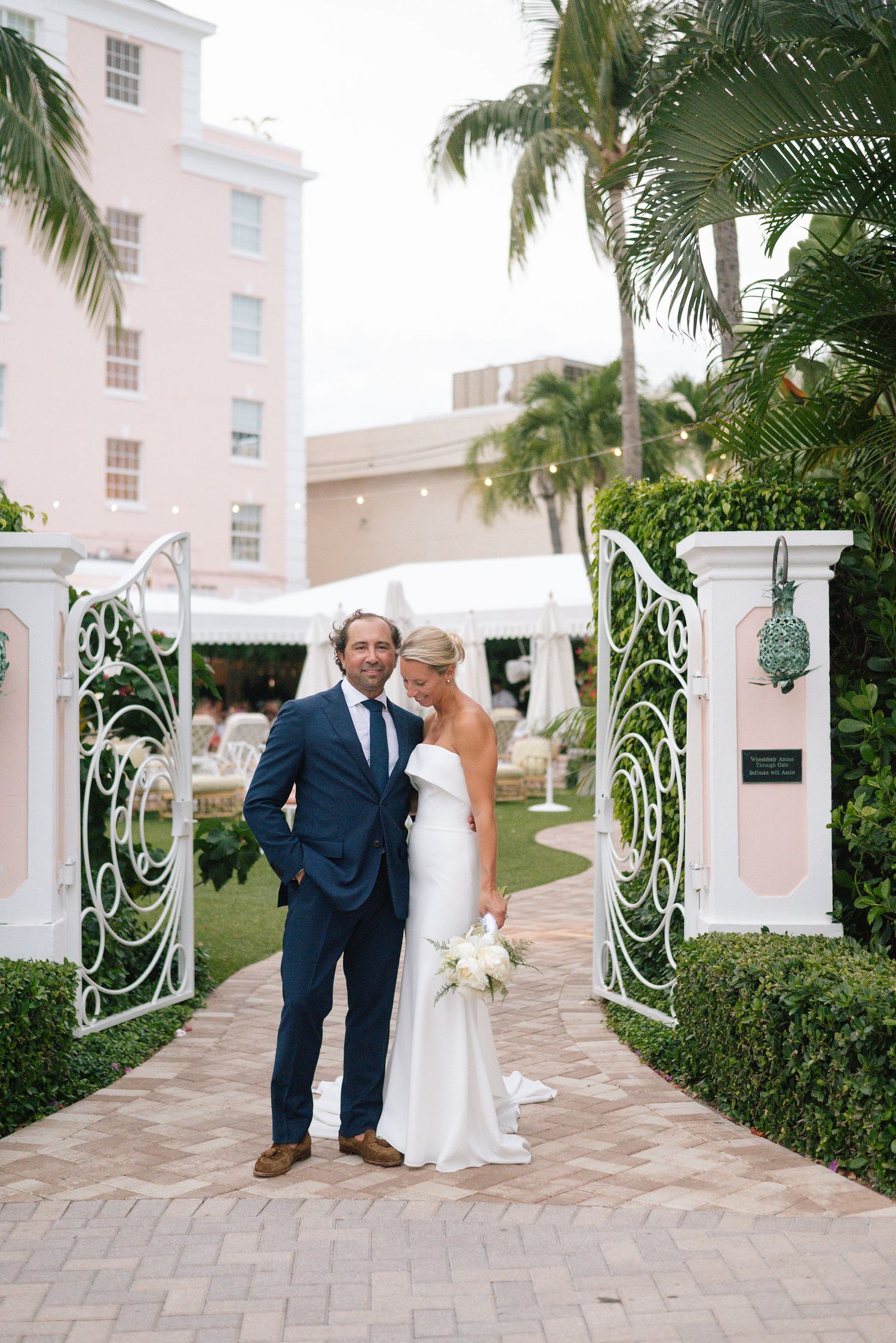 Wedding couple shares a romantic moment on a palm-lined pathway at a pink hotel entrance.