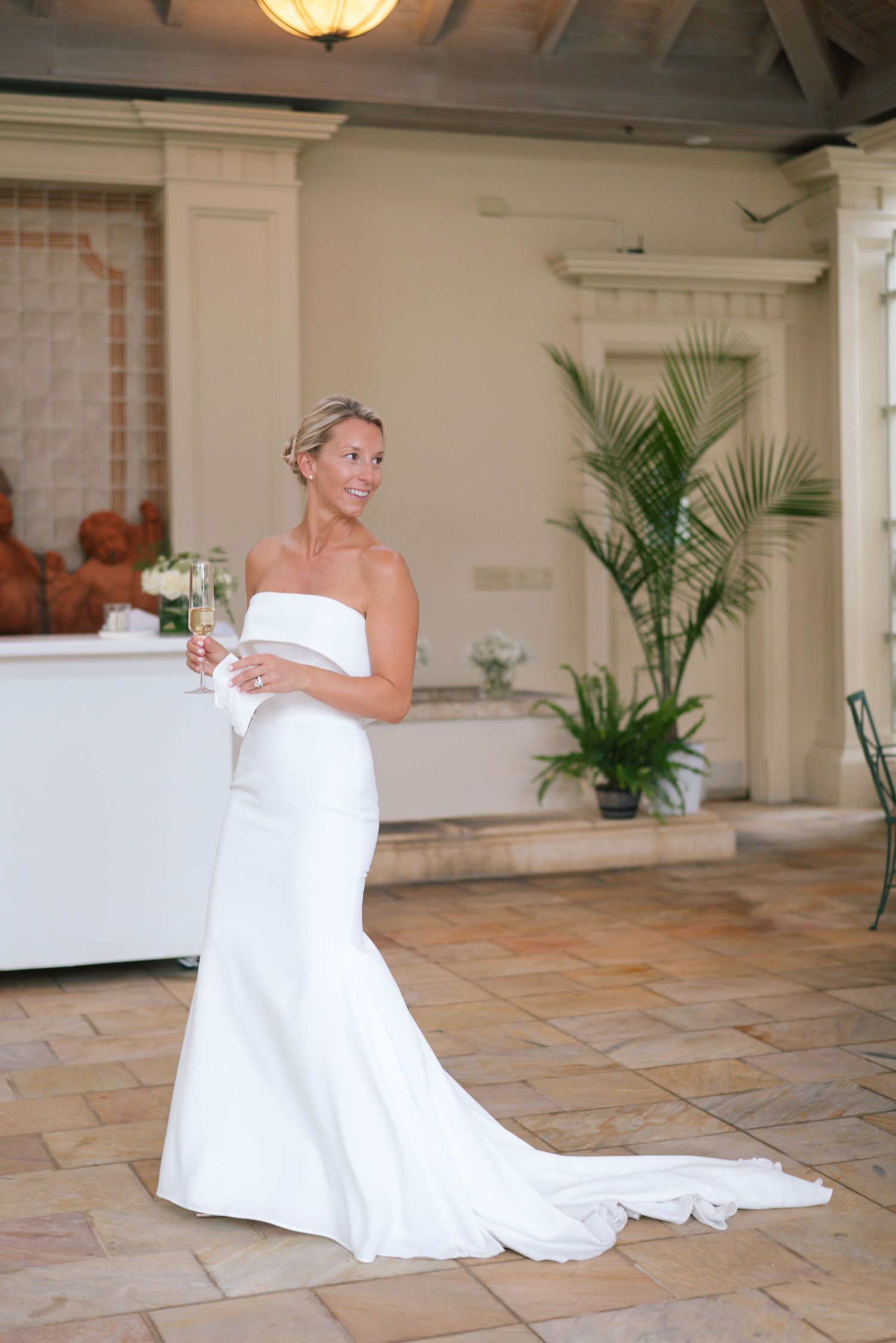 Elegant bride in fitted white gown holding champagne in a conservatory setting.