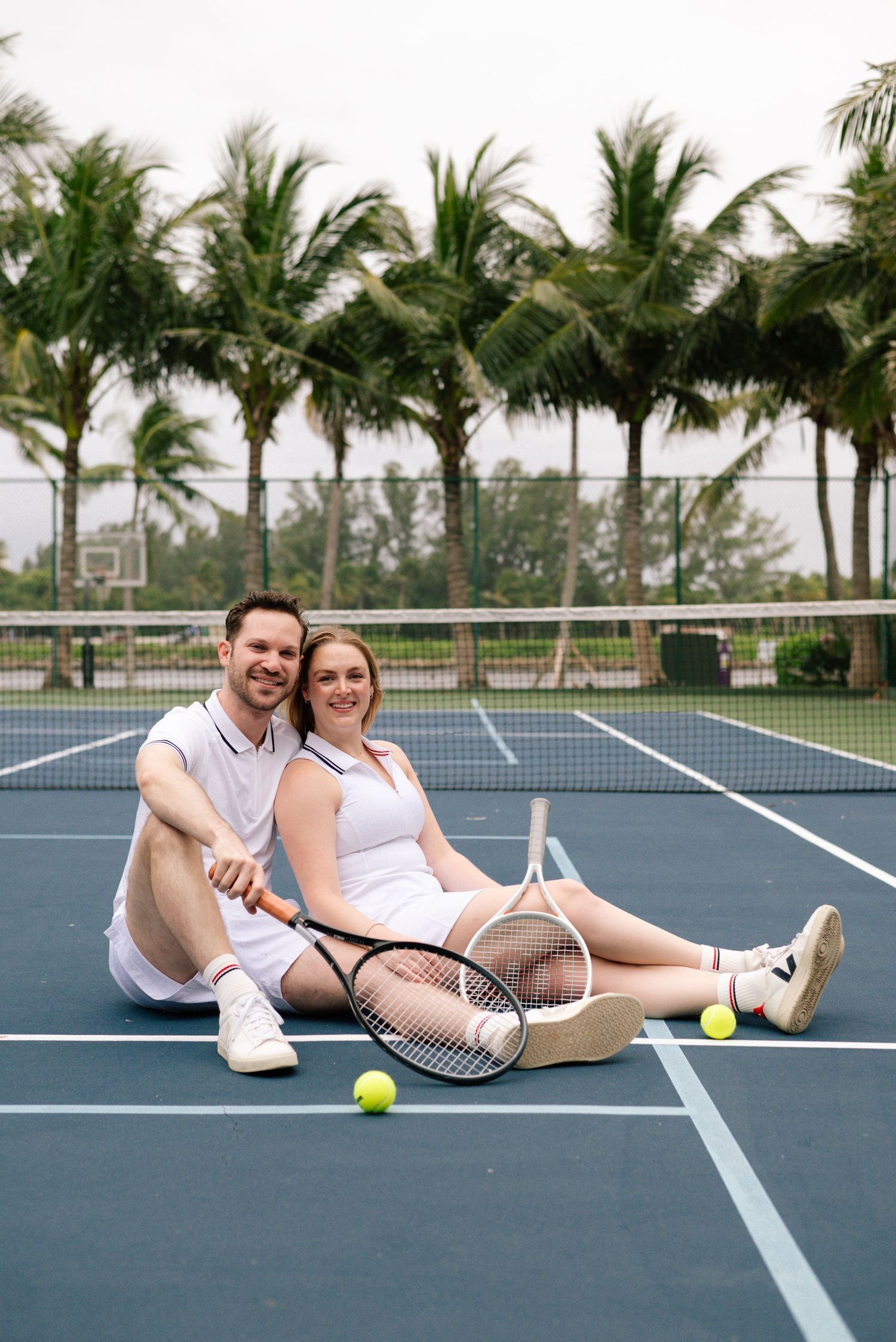 A couple sits together on a blue tennis court with rackets and tennis balls around them.