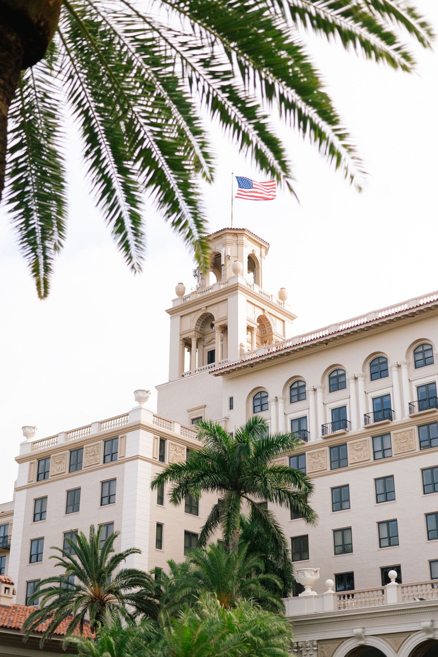 Elegant Mediterranean-style resort building with white facade and palm trees under blue sky, featuring American flag.