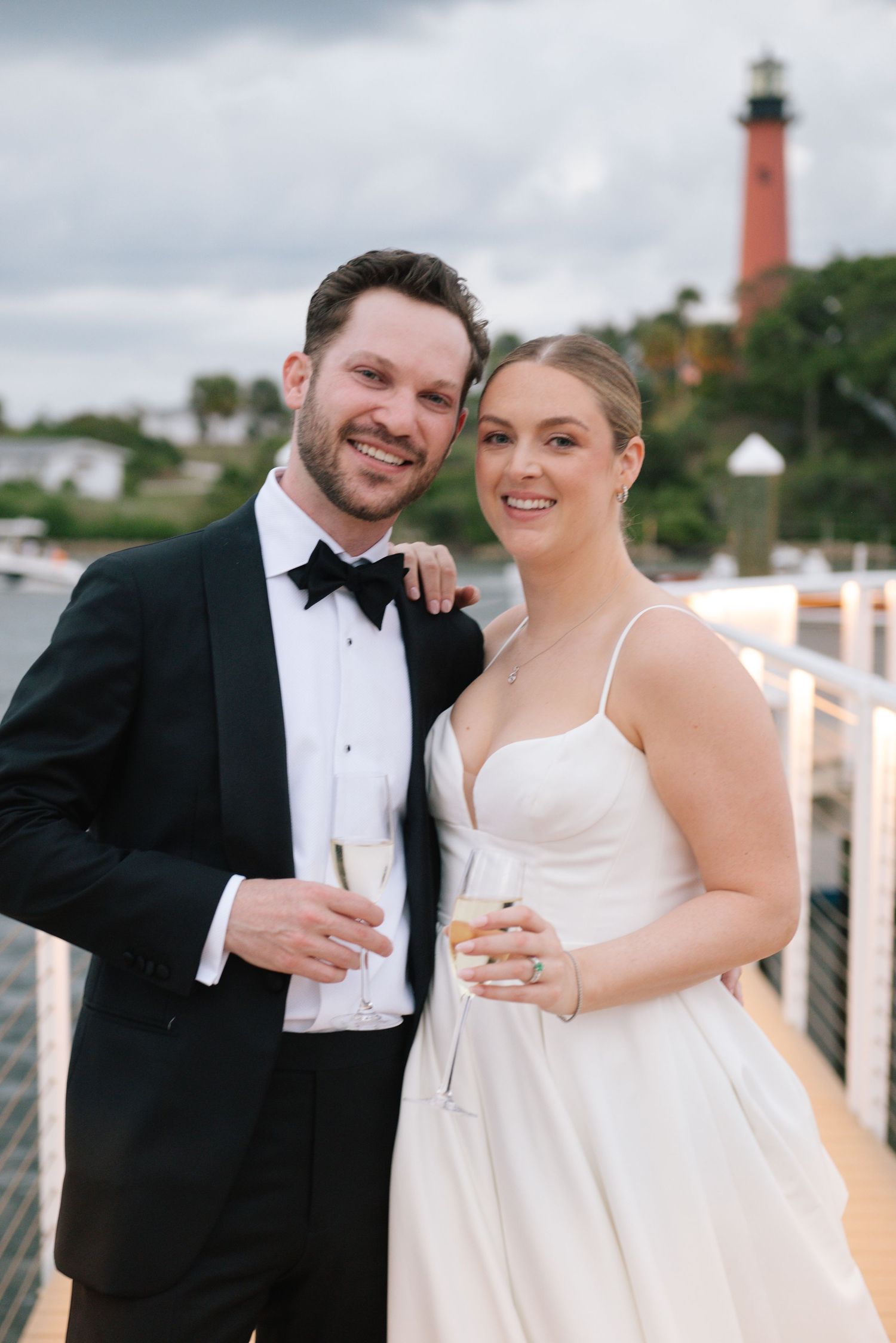 A wedding couple poses on a boardwalk with a historic lighthouse visible in the background.