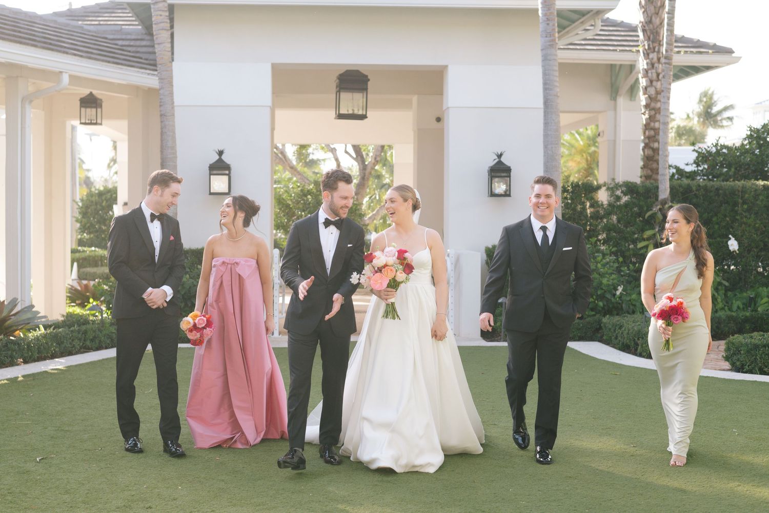 Wedding party in formal attire walking together on manicured lawn outside elegant venue.