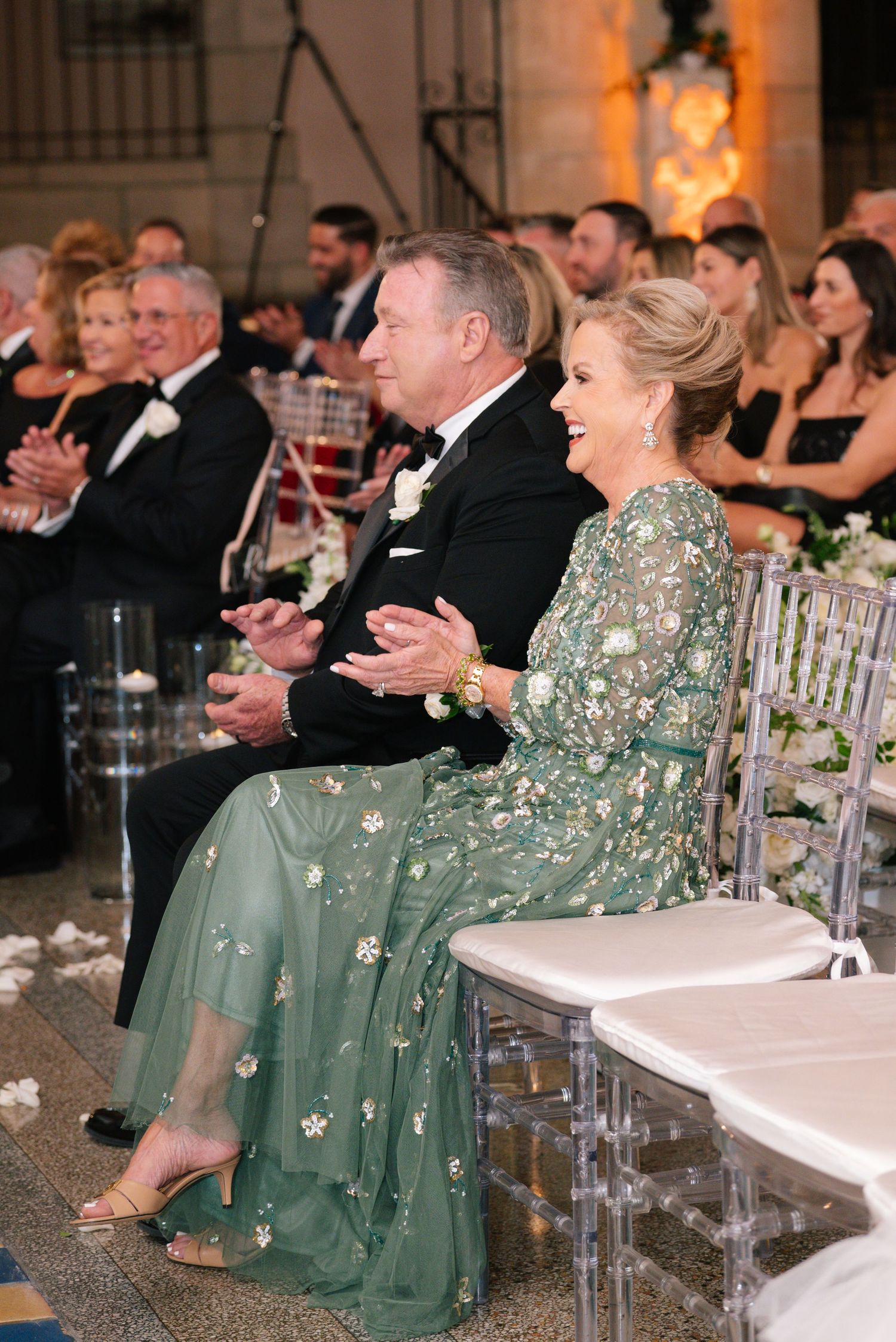 Mother of bride in green floral gown and father in tuxedo applaud during elegant wedding ceremony.