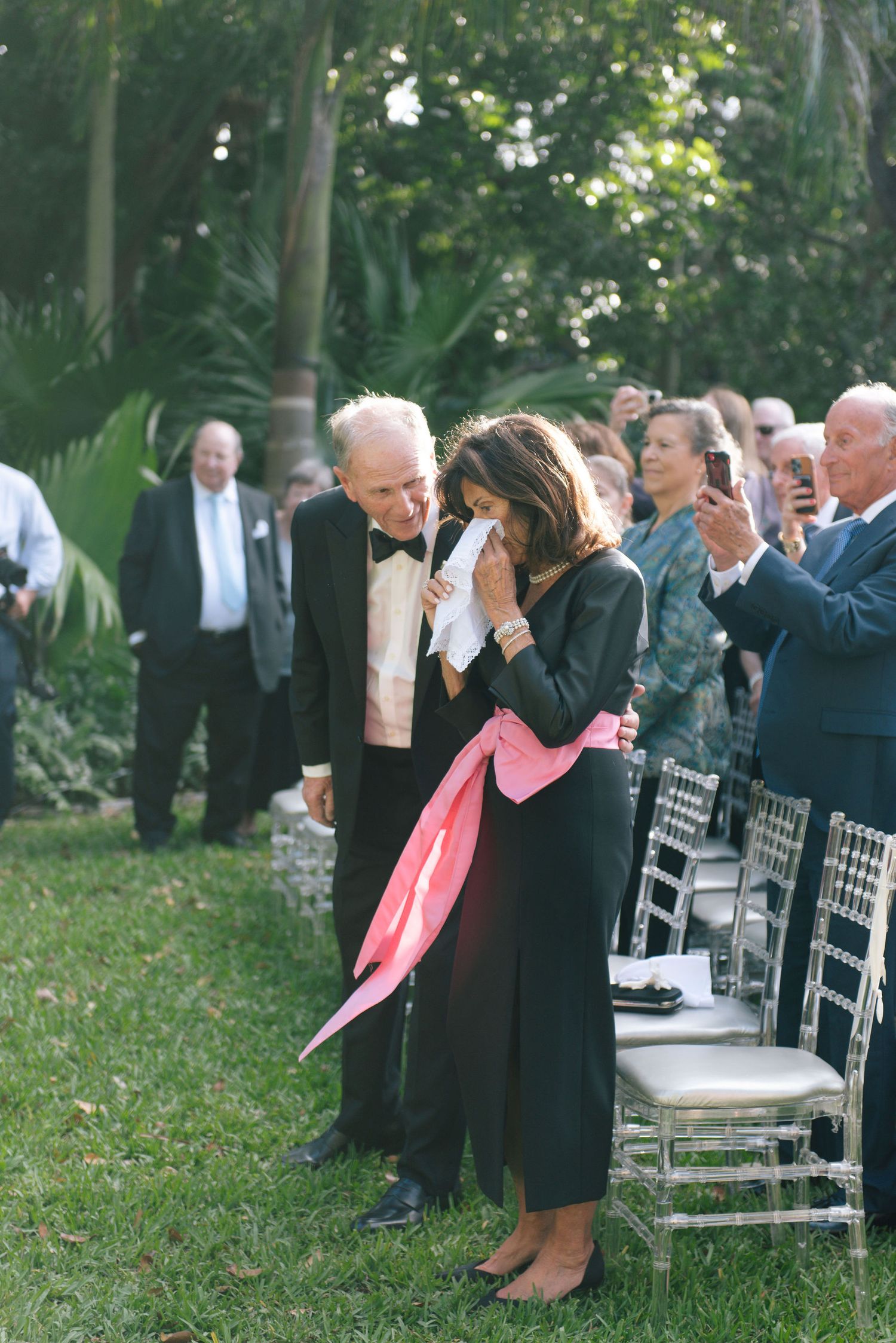 Wedding guests seated on white chairs in an outdoor garden setting with palm trees.
