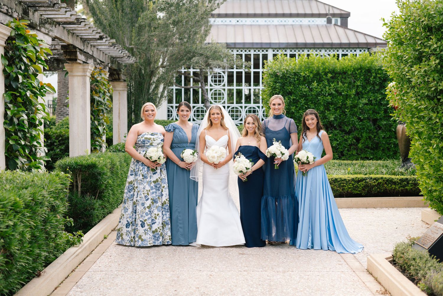 A group in blue dresses and formal attire poses on stone steps surrounded by lush garden landscaping.